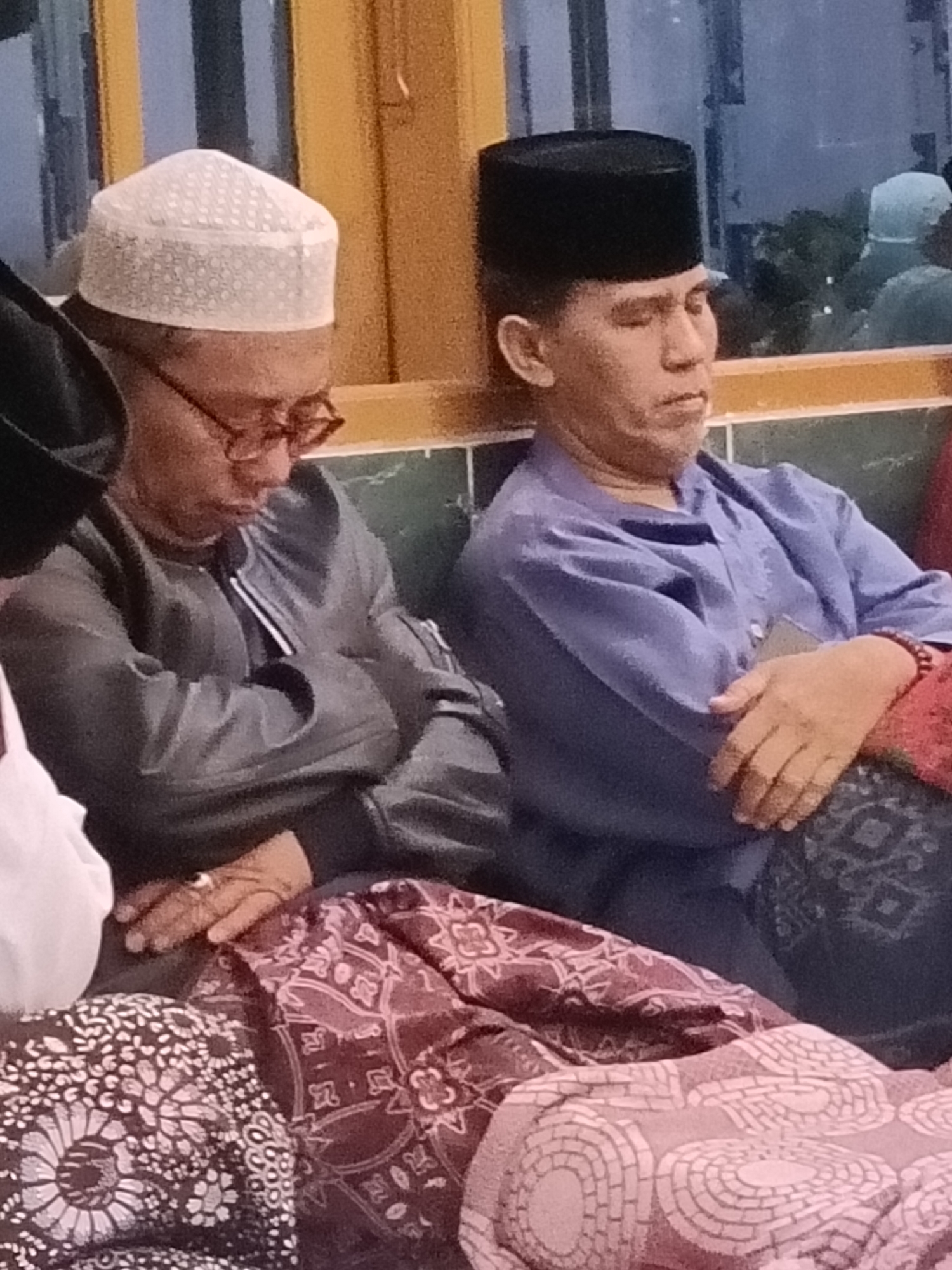 The image shows two men sitting indoors, possibly in a mosque or prayer room in Kediri City, Indonesia. The man on the left wears a white patterned cap and glasses, and a grey jacket. He is looking down, with his hands resting on his lap. The man on the right wears a black cap and a blue shirt, with his eyes closed. His arms are crossed, and he is wearing a bracelet on his right wrist. Both men appear to be resting or sleeping. In the background, there is a window with reflections of trees and sky. The floor is covered with patterned rugs.