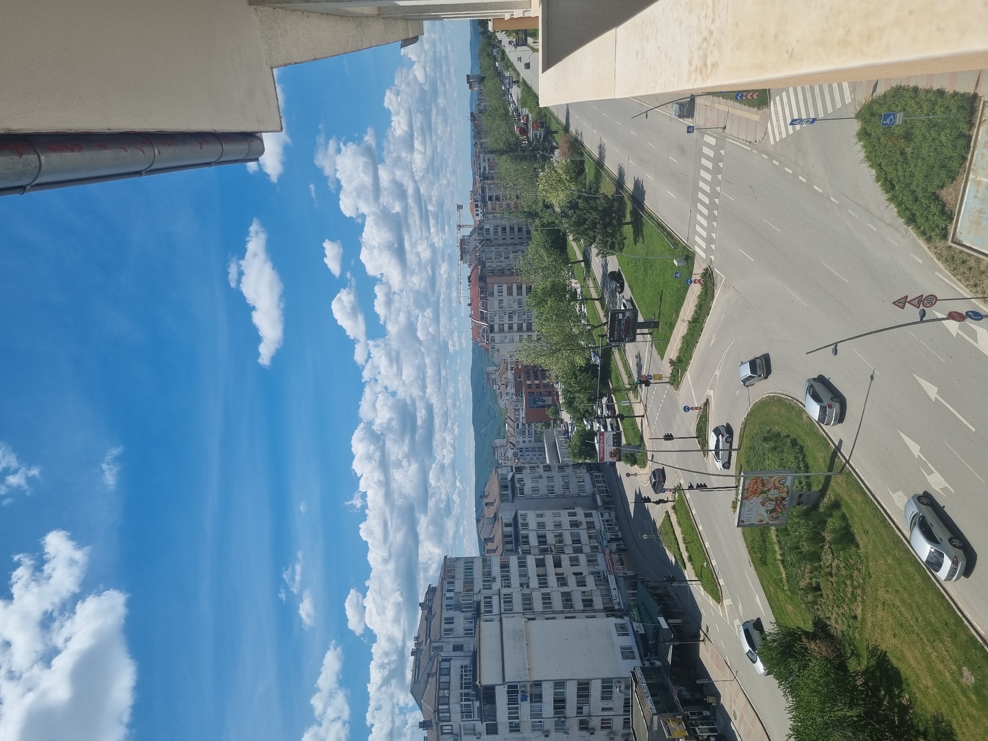 This is an elevated, urban view in Veliko Tarnovo, Bulgaria, showing a street intersection. The sky is largely blue with scattered white clouds. The street has cars on it, including silver and white vehicles, and green grassy medians. Buildings line the street, ranging from smaller structures to multi-story apartment buildings. A billboard is visible on one of the medians. Road markings and pedestrian crossings are present. The view is partially framed by architectural elements, likely a balcony.