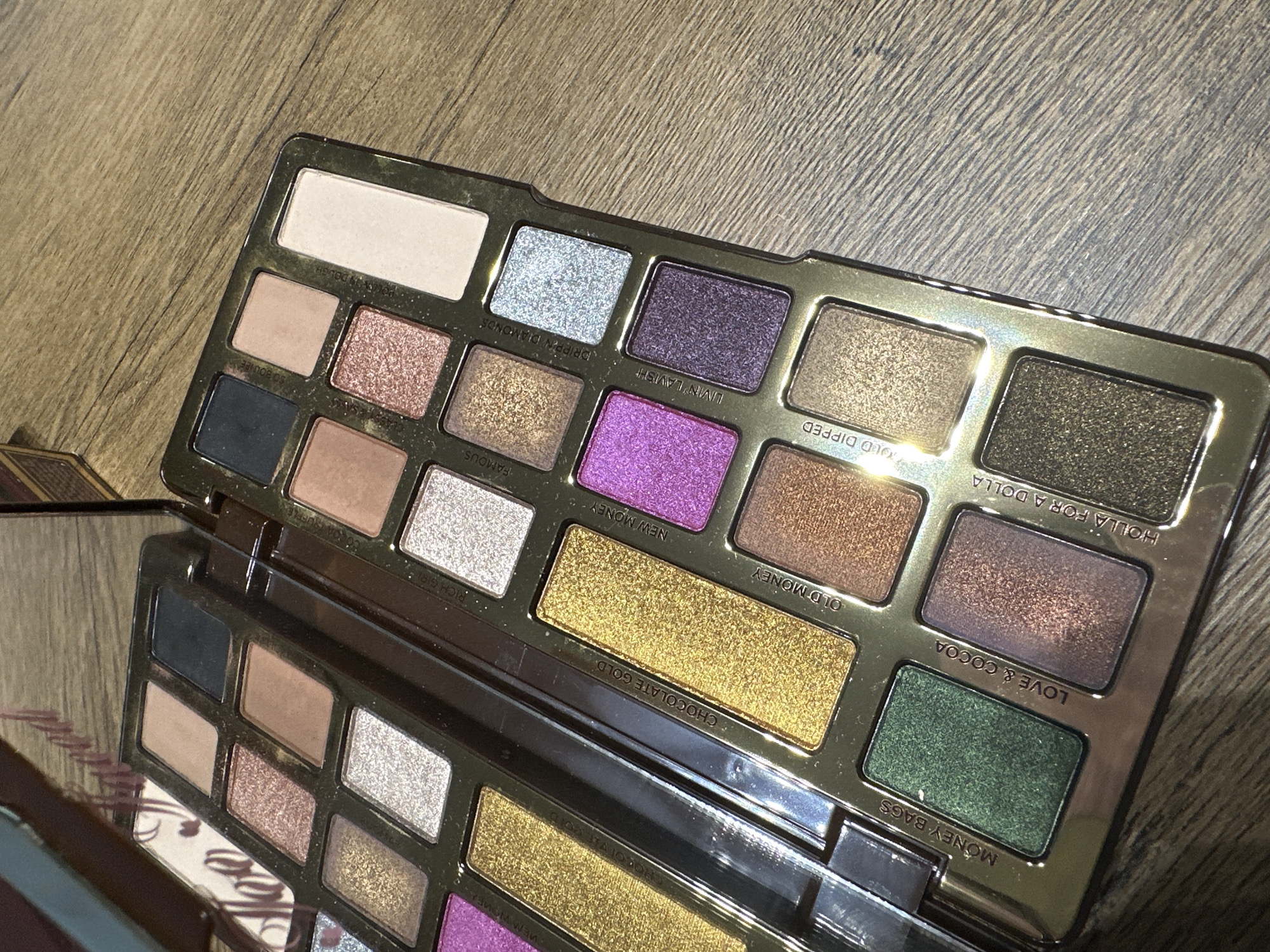 A makeup palette is on a wooden surface. The palette, primarily gold-toned, features a variety of colors and textures in rectangular pans. A mirror on the side reflects a partial view of the palette. There are no people visible in the image, nor any information about this being taken in Plovdiv, Bulgaria.
