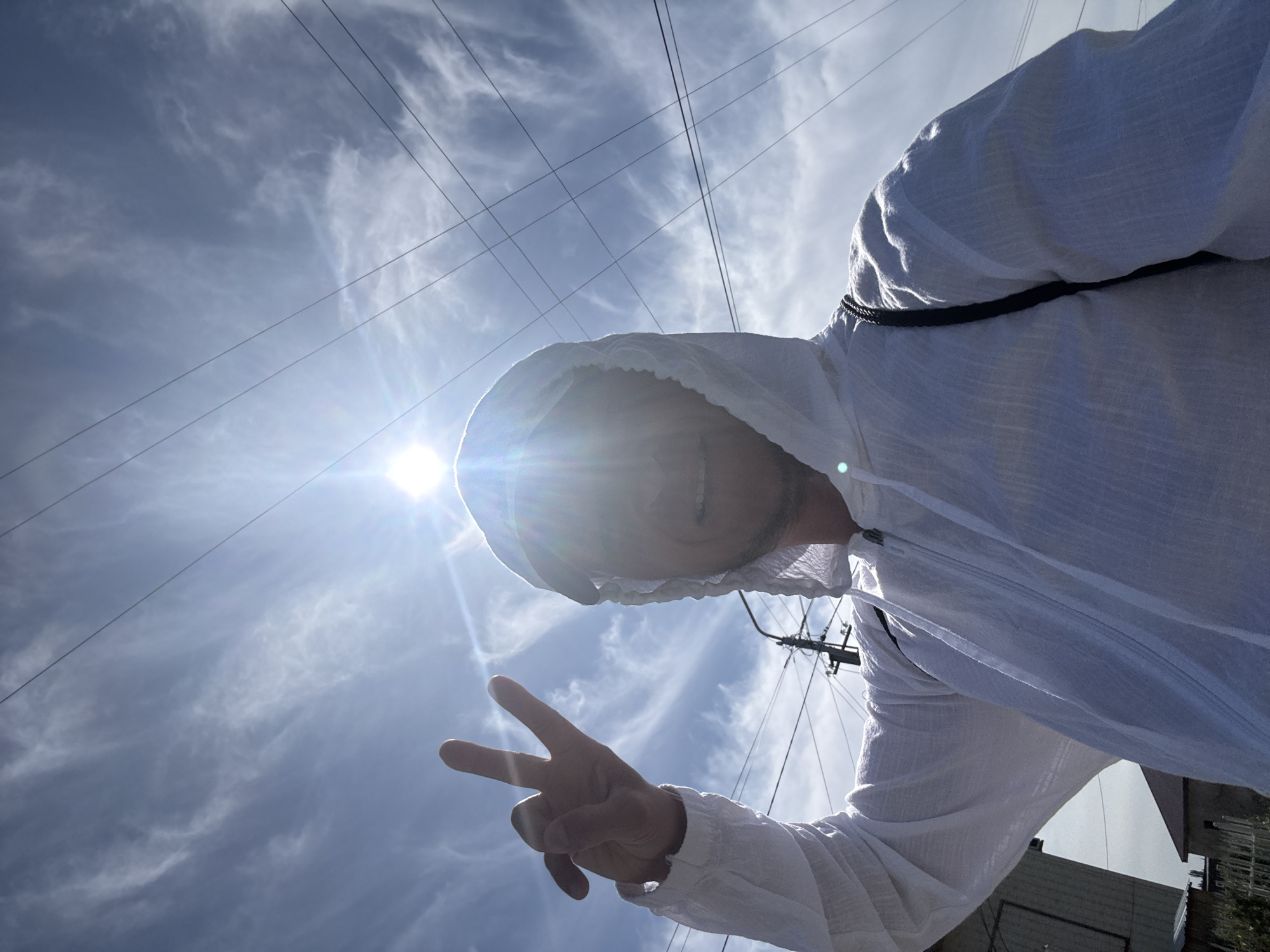 A person wearing a white hooded jacket and holding up a peace sign is pictured outdoors. The sun is bright and partially obscures the face of the person. In the background, there are power lines against a blue sky with wispy clouds. The person appears to be on a street with buildings visible to the left. Based on the context provided, the location is the city of Zharbulak, Kazakhstan.