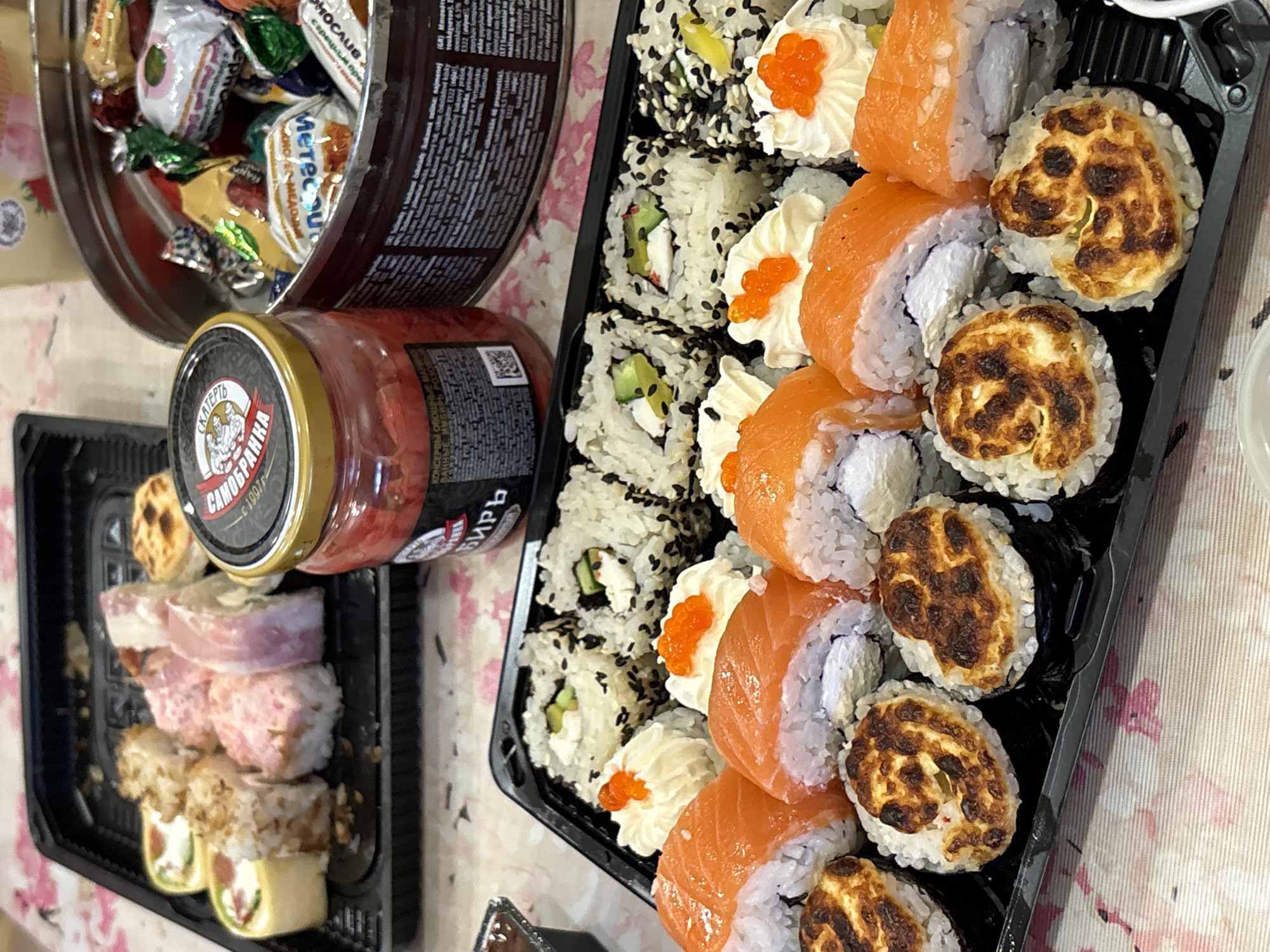 This image shows a table setting with various food items. There's a round metal tin filled with candies, a jar of what appears to be preserved vegetables, and two black plastic trays filled with different types of sushi. The table has a floral patterned tablecloth. The jar is labeled "SKATERT САМОБРАНКА с 1997г". It includes Russian text. The location in the city of Berezniki, Russia, is not explicitly stated.