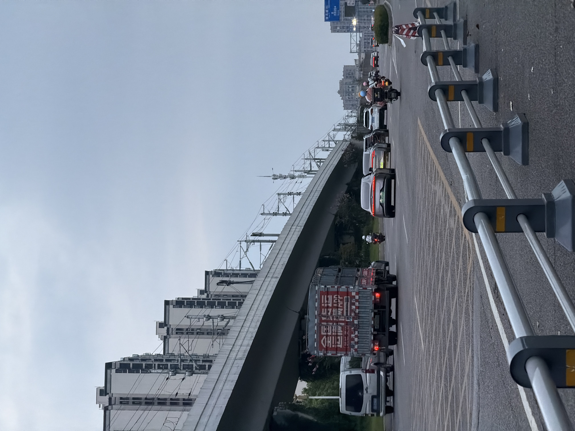 An overcast day in Jiangjin, China, features a multi-lane road with several vehicles. In the foreground, a silver van and a red truck with Chinese characters on its side are traveling in the same direction. The truck is carrying goods. Behind them, a silver sedan and several other cars are visible. A motorcyclist wearing a blue helmet and a passenger are also on the road, as are other vehicles further in the distance. Elevated train tracks run overhead, supported by concrete pillars, with numerous buildings lining the tracks. The sky is a light gray, indicating cloudy weather. The road has clear lane markings and a metal barrier separates lanes of traffic. A traffic sign is visible on the right side of the road, though its text is unreadable.