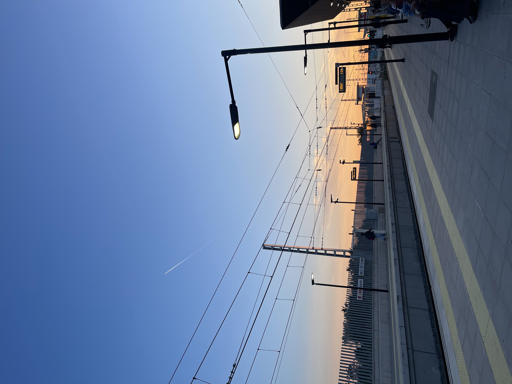 The image depicts the Paiporta metro station in Paiporta, Spain, at sunset. The foreground shows the platform, with a person walking along it. Overhead electrical lines stretch across the frame, juxtaposed against a gradient sky that transitions from blue to orange. Street lights illuminate the platform. In the distance, more people can be seen waiting. An airplane contrail cuts across the upper left sky.