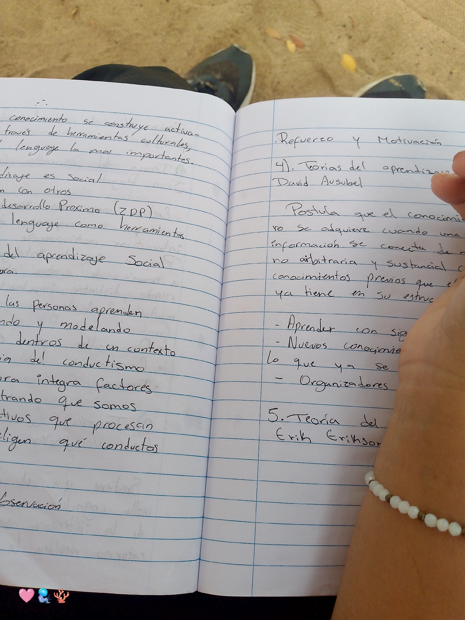 The image shows an open notebook on a lined page. The left page contains handwritten notes in Spanish about knowledge construction, cultural tools, language, social learning, and behavior. The right page contains notes about reinforcement, motivation, and theories of learning by David Ausubel, including postulates about acquiring knowledge and connecting information to previous knowledge. It also mentions learning new information, organizers, and Erik Erikson's theory. The edge of a person's arm is visible, adorned with a white beaded bracelet. The notebook is placed on top of a person's legs. In the background, there are a couple of dark-colored shoes. Although the prompt requests this image to be located in Pedro González, Venezuela, the image alone does not confirm this location.