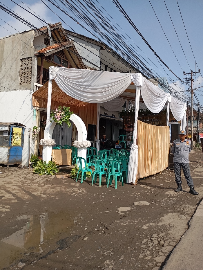 The image depicts an outdoor event setup on a dirt road in Cipatik, Indonesia, under a clear blue sky. A temporary canopy, draped with white and gold fabric and supported by white pillars adorned with artificial flowers, covers an area furnished with multiple rows of empty green plastic chairs. A vertical banner visible within the canopy displays the text "Walimatussafar Haji & Khitan" in Indonesian. A man in a grey uniform, including a cap, dark pants, and black boots, stands on the right side of the frame, facing the canopy. Several indistinct individuals are visible seated or standing further back under the canopy. The unpaved ground in the foreground is uneven, showing dirt, cracks, and a large puddle reflecting the sky. Numerous black utility wires crisscross overhead. In the background, two multi-story buildings with aged facades are visible, and a blue metal kiosk or bin is present on the far left.