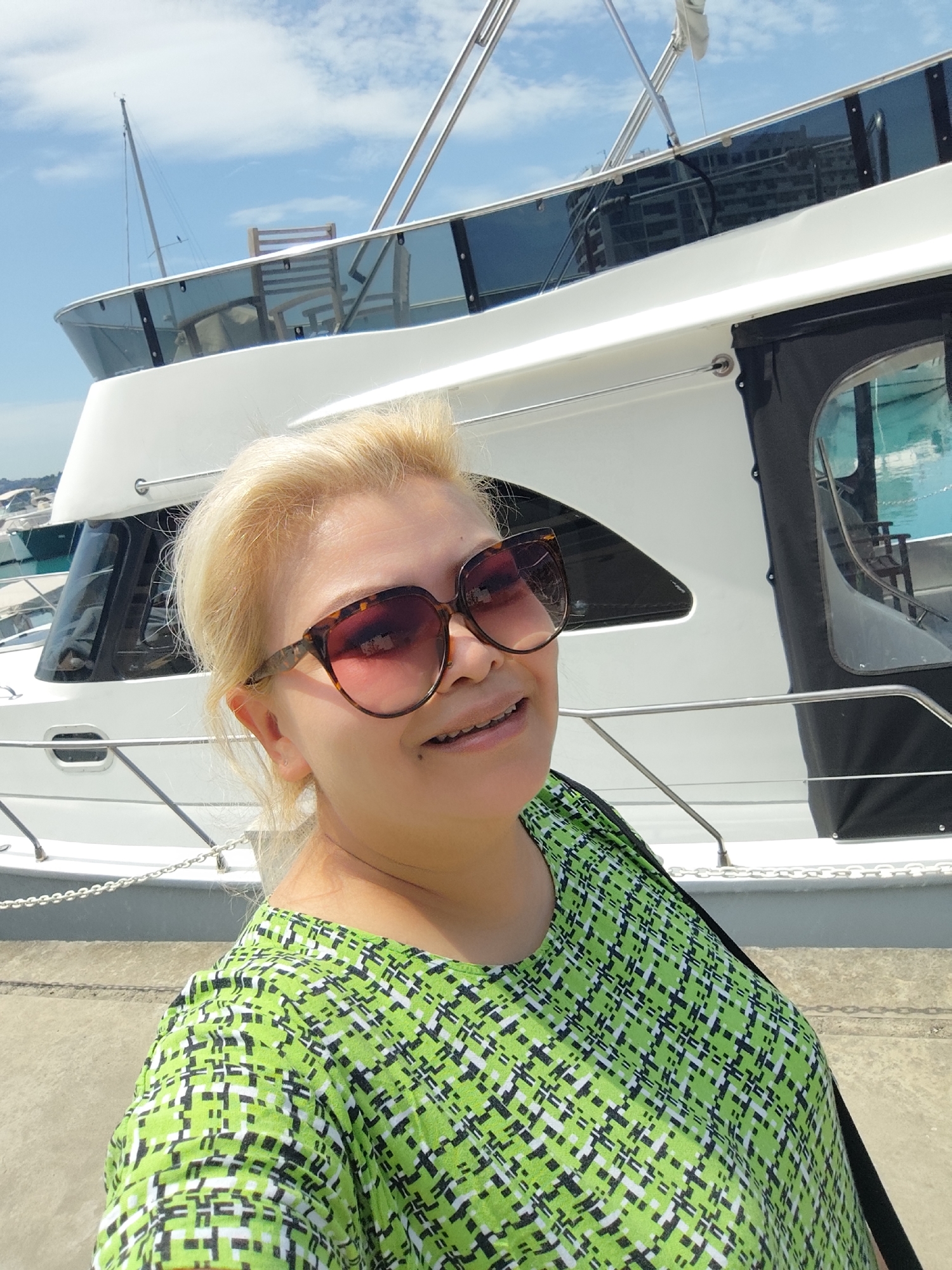 A full-color, eye-level selfie taken on a bright, sunny day features a woman with blonde hair, smiling at the camera. She wears large, tortoise-shell sunglasses and a vibrant green top with a black and white geometric pattern. A black strap from a bag is visible over her right shoulder. Behind her, a prominent white yacht with dark tinted windows is docked at a marina. Reflections of distant buildings and the sky are visible in the boat's windows. In the background, under a blue sky with scattered clouds, other boats and masts are visible. The woman is standing on a concrete dock or pier. Her smile conveys a happy and relaxed mood, suggesting she is enjoying her time. The bright sunlight and clear sky indicate it is daytime. No text is visible in the image. The setting is consistent with a marina in Sarıyer, Turkey.