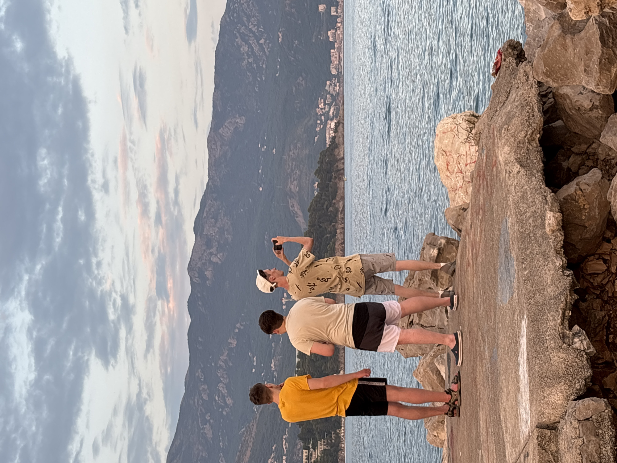 Three young men stand on a rocky outcrop overlooking the Adriatic Sea in Budva, Montenegro. The man on the left wears a yellow t-shirt and black shorts. The middle man is dressed in a cream-colored t-shirt and black and white shorts. The man on the right, wearing a patterned t-shirt, a white cap, and grey shorts, is holding a smartphone up to take a photograph. The sea is calm, and in the background, the hilly coastline of Budva is visible, dotted with buildings and covered in lush greenery. The sky above is a soft blend of grey and pink clouds, suggesting either dawn or dusk. The rocky shore is composed of large, weathered stones.