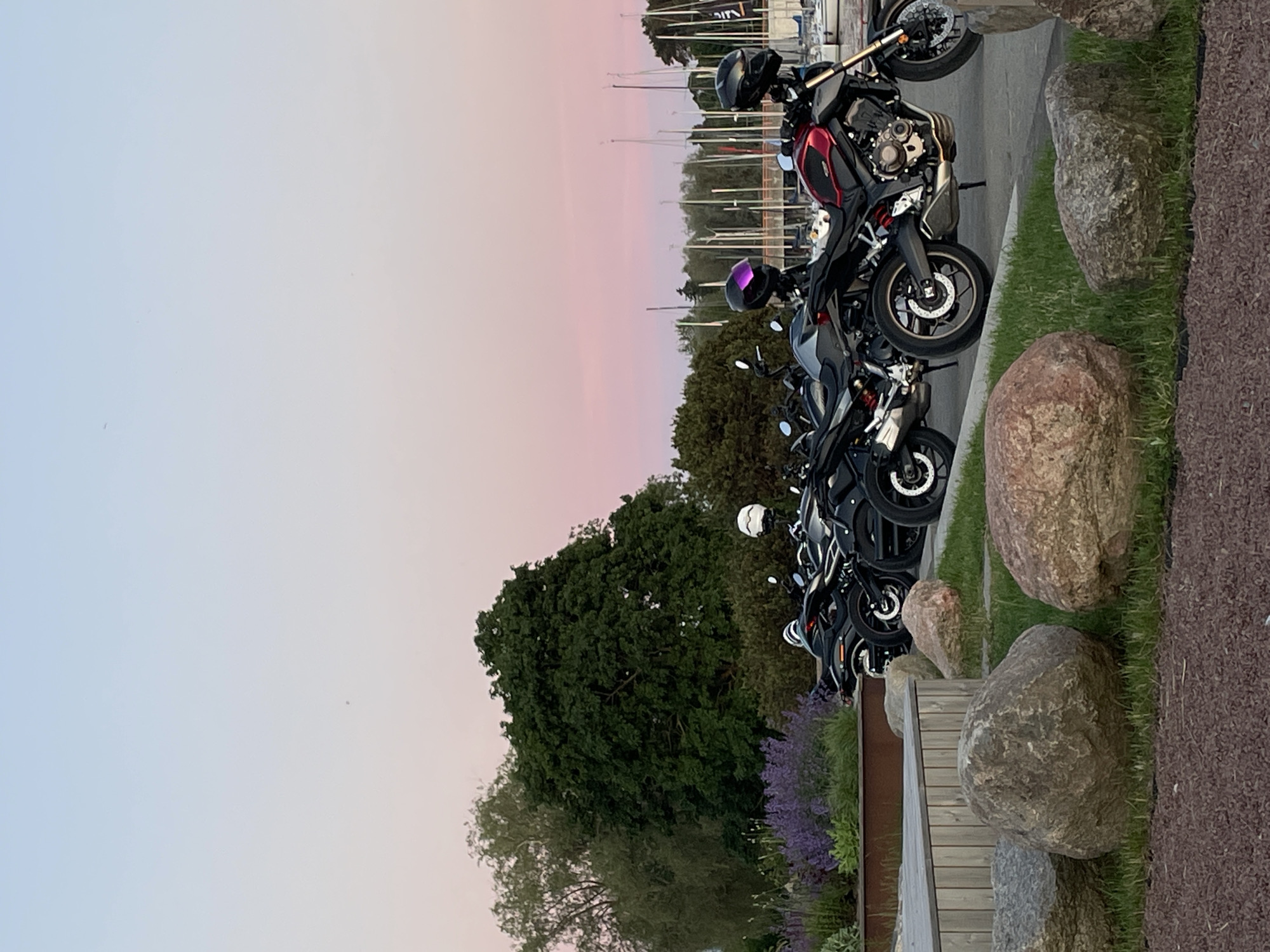 The image depicts a row of parked motorcycles in the foreground, with a serene harbor and silhouetted sailboats in the background. The scene is set against a pastel-colored sky, transitioning from a soft pink to a pale blue, indicating either sunrise or sunset. Large, weathered rocks and lush green grass occupy the immediate foreground, adding a natural element to the urban waterfront setting. The motorcycles are diverse in color and design, with one prominently featuring a red and black color scheme. Helmets are visible on some of the bikes, suggesting recent use or preparation for a ride. The overall atmosphere is tranquil, highlighting the juxtaposition of modern machinery with the natural beauty of the coastal environment. The location is the Tallinn harbor area, specifically near the Seaplane Harbour Museum, a popular waterfront district in Tallinn, Estonia.