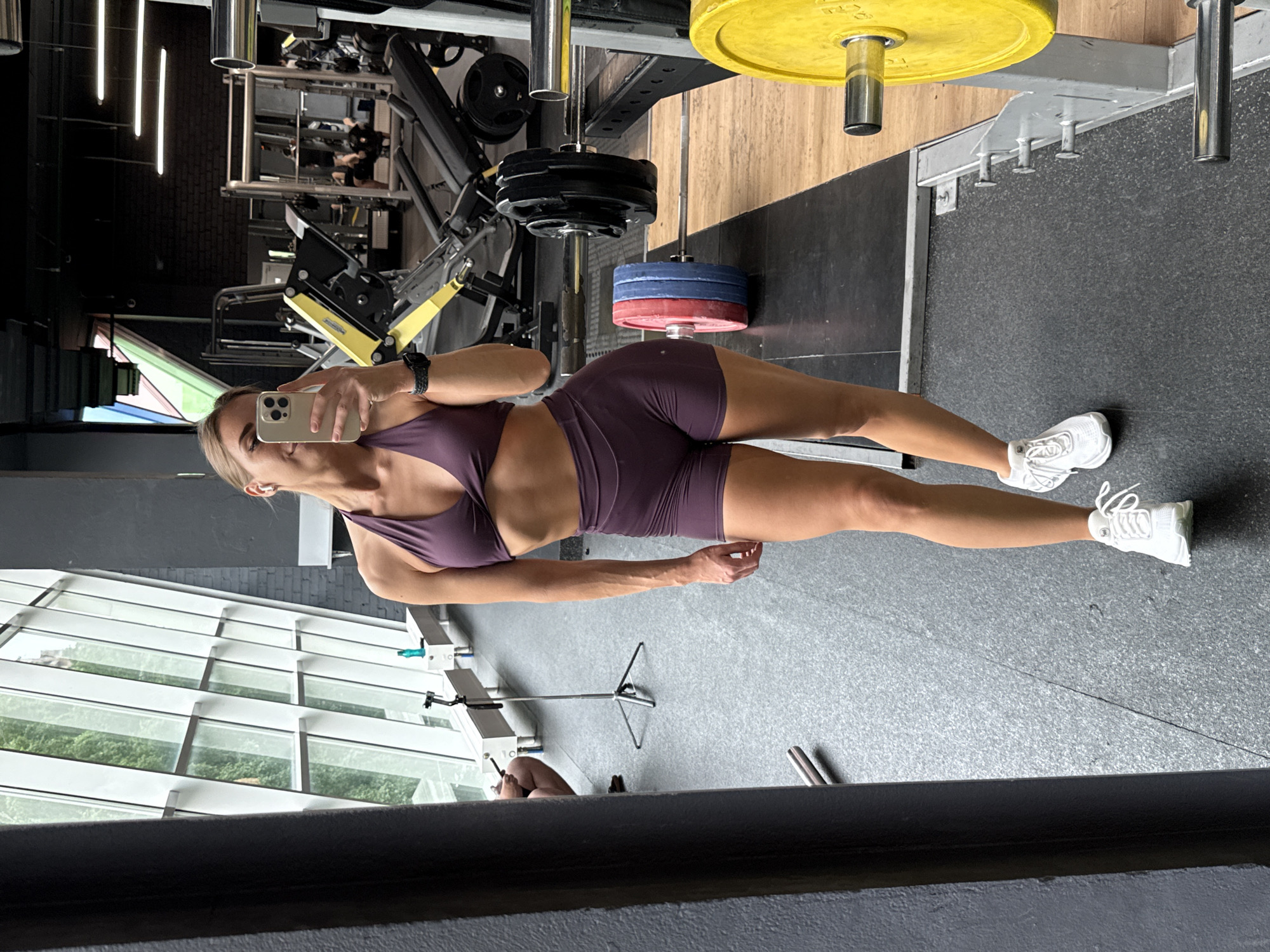 A young woman with blonde hair pulled back is taking a mirror selfie in a gym. She is wearing a dark purple sports bra and matching shorts, along with white athletic shoes. The gym appears to be modern with large windows, weightlifting equipment, and rubberized flooring. A tripod with a phone is visible on the floor near the windows. The woman is standing in front of a large mirror, capturing her reflection. The scene is consistent with a gym located in Saint Petersburg, Russia.