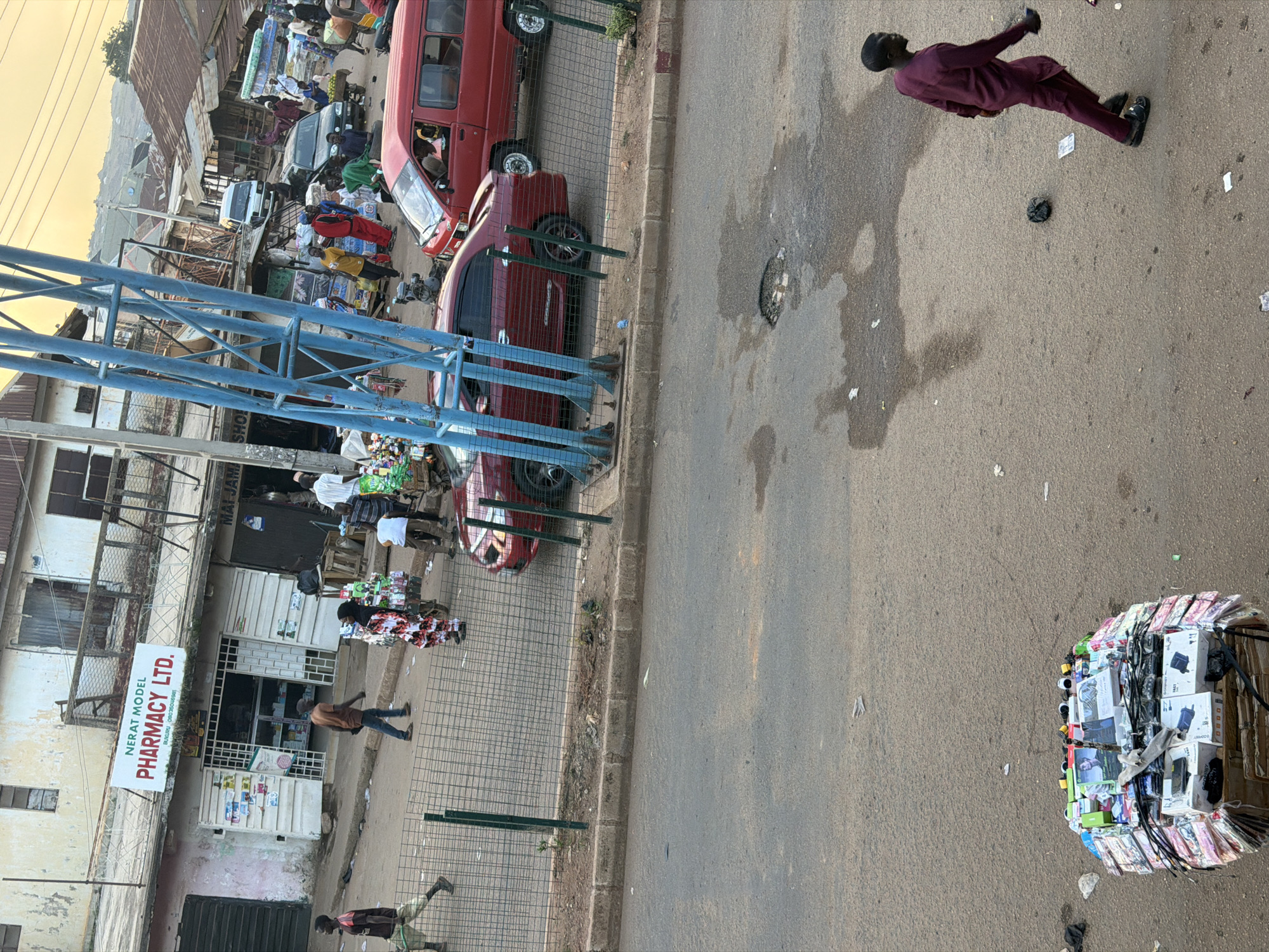 The image shows a street scene in Bukuru, Nigeria. In the foreground, a man in a maroon robe walks across a paved road. A large pile of boxed goods is on the left side of the road. In the middle ground, a red sedan is parked. A red van is behind it. A wire fence separates the road from a sidewalk where several people are walking. Behind the sidewalk is a row of shops. One shop has a sign that reads "NERAT MODEL PHARMACY LTD." The shops are on a street with buildings in the background. The sky is bright and overcast.