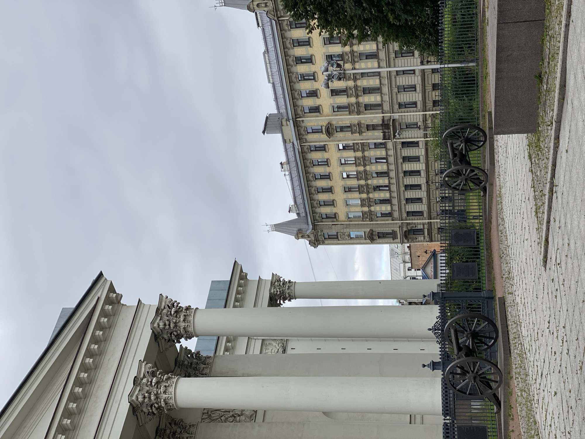 The image depicts an outdoor scene in Saint Petersburg, Russia, likely in front of a grand neoclassical building with white columns. In the foreground, two vintage cannons on wheeled carriages are positioned behind a dark metal fence. The ground is paved with light-colored square stones, interspersed with small patches of green grass.
Beyond the fence, an ornate, pale yellow apartment building with numerous windows and classical architectural details dominates the middle ground. A tall, dark-green tree is visible to the right of this building. The sky above is overcast and a uniform gray, suggesting cloudy weather. The overall impression is one of historical significance and architectural grandeur.