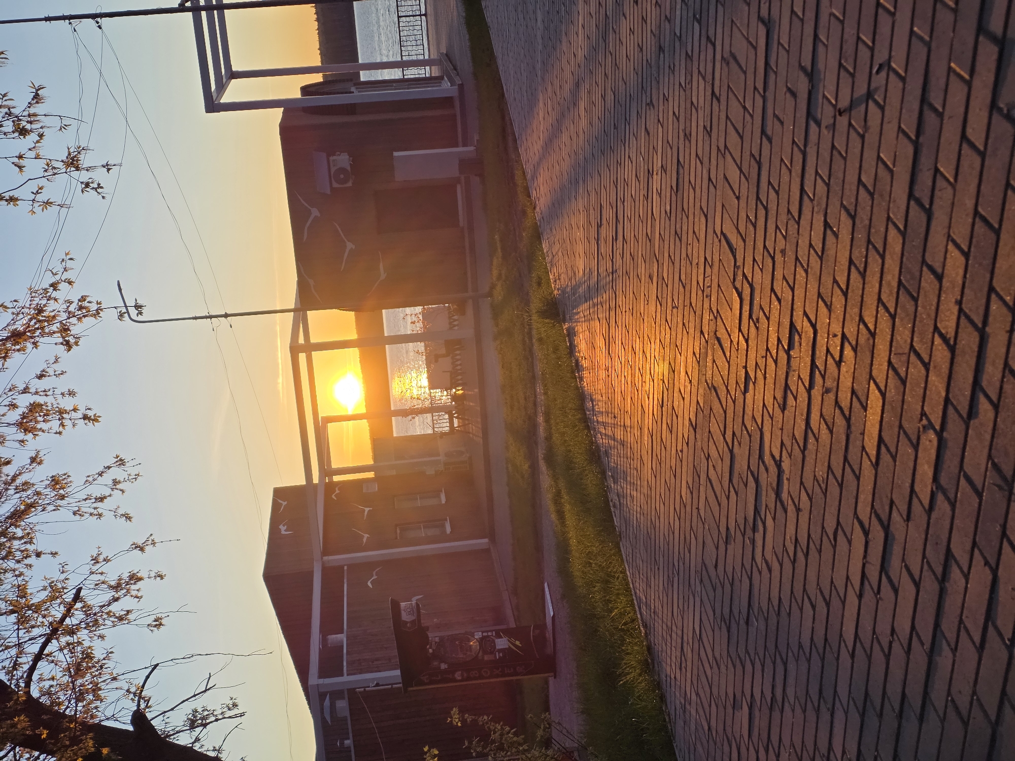 The image depicts a brick-paved walkway alongside a grassy strip, with buildings visible in the background. The sun is setting, creating a bright, golden glare in the center of the frame. The location is Chaikovsky, Russia. The image is rotated counter-clockwise and needs to be properly oriented in order to view it correctly. A tree with sparse leaves is visible in the lower-left corner. The buildings appear to be recreational or commercial structures, and one has a sign visible in front of it. The sun is reflecting off the water in the distance, which appears to be a lake or river.