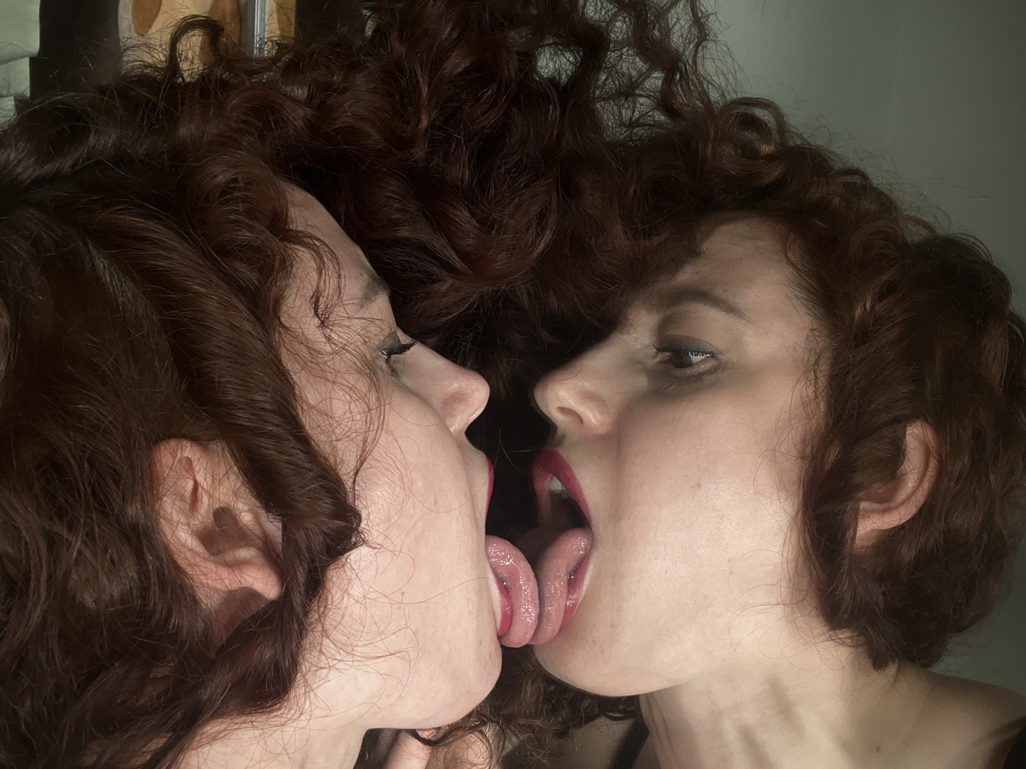 The image shows a close-up of two female faces with dark red curly hair touching each other. Their lips are open, and their tongues are touching. Both women have red lipstick. The background is blurred and undefined, but it appears to be a room with light-colored walls. There are no visible details that suggest the location is in Saint Petersburg, Russia.