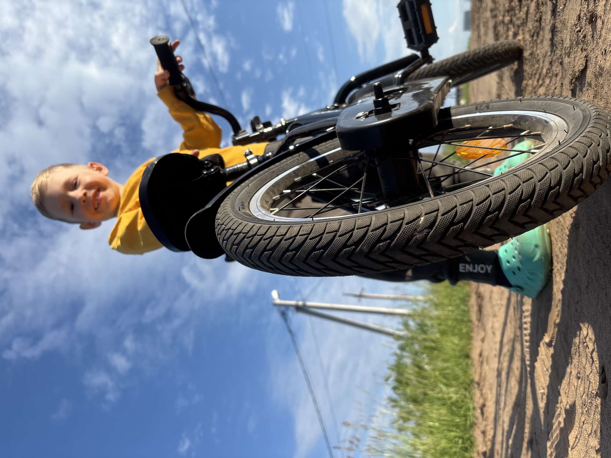 A young boy, smiling widely, is captured in a low-angle shot from the perspective of his bicycle. He is wearing a bright yellow hoodie. The bicycle's front wheel is prominently in the foreground, with its textured tire dominating the lower half of the frame. The boy's hand is on the handlebars, and his foot, clad in a turquoise clog, rests near the pedals. The bicycle frame is black.
The scene unfolds outdoors under a bright blue sky dotted with white, fluffy clouds. Sunlight suggests it is daytime. The ground appears to be a sandy or dirt path, with patches of green grass visible on the right side of the image. In the background, a utility pole and power lines are discernible, hinting at a somewhat rural or suburban setting. The overall mood is cheerful and energetic, with the boy's expression and the bright lighting contributing to a sense of fun and freedom. The word "ENJOY" is visible on a black component of the bicycle, possibly a pedal or part of the frame, reinforcing the positive sentiment. The image is oriented vertically, with the subjects appearing upside down from a conventional perspective.