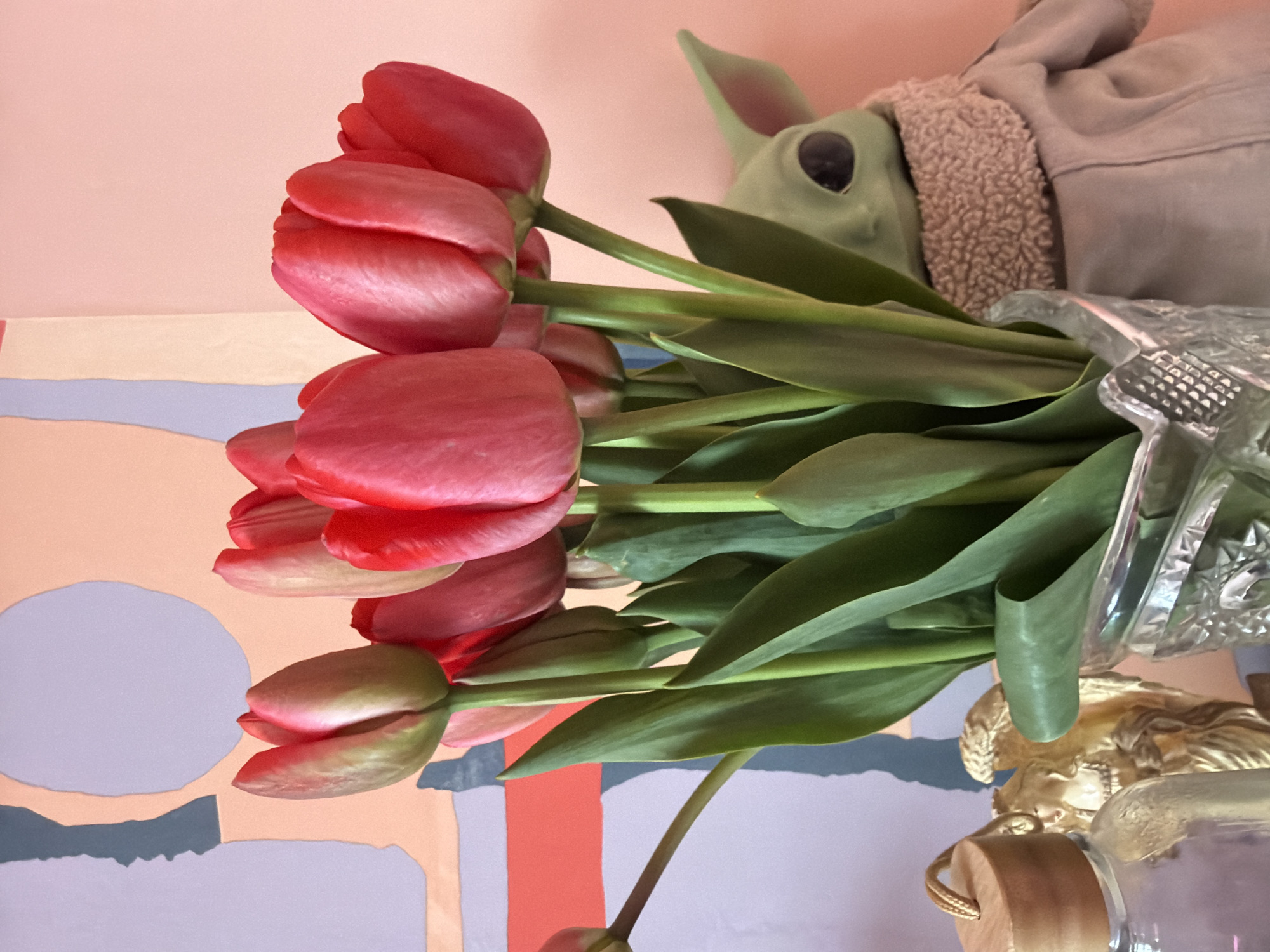 A vase of red tulips sits on a surface next to a Grogu (Baby Yoda) figurine wearing a suede jacket with a fleece collar. Under the vase is a gold-painted figure and a glass jar with a wooden lid. The background features a wall with pastel shades of pink, purple, grey, and orange, patterned in abstract shapes including a circle. The location is not specified but cannot be independently confirmed as Chaikovsky, Russia.