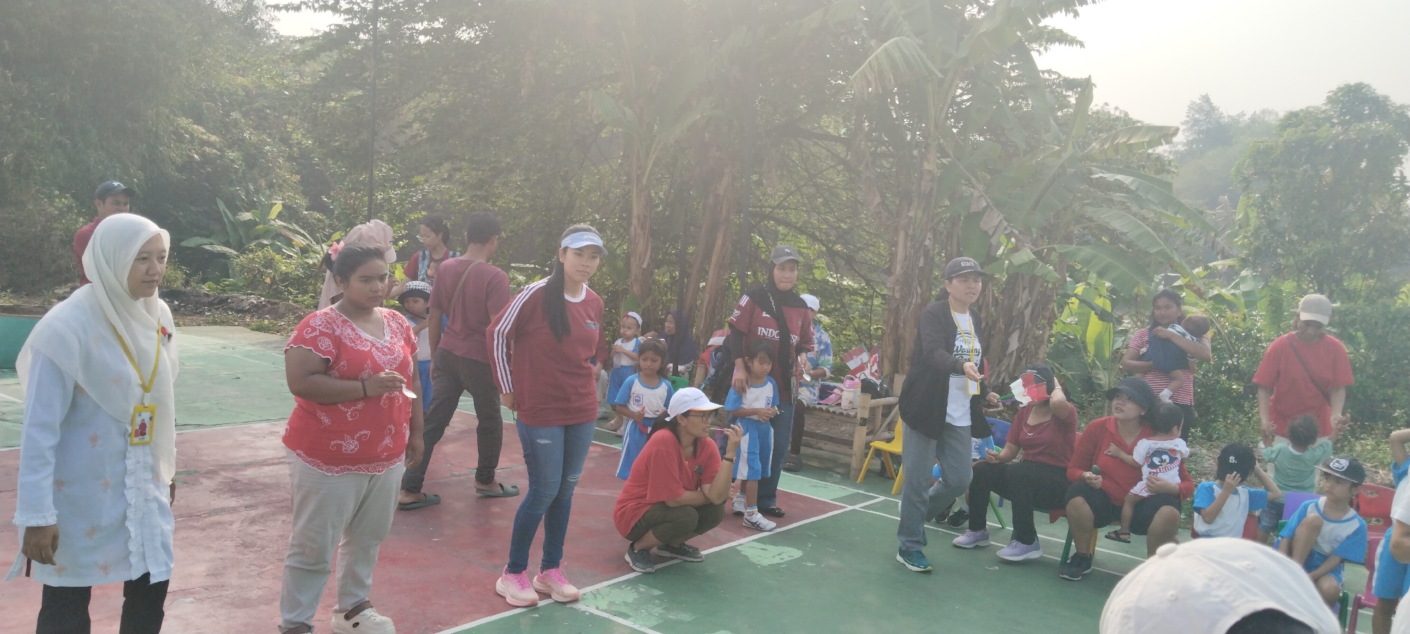 The image depicts an outdoor gathering, possibly a school event or community activity, set against a backdrop of lush greenery, suggesting a location in or near a rural or semi-rural area of Tabanan, Indonesia. In the foreground, a group of people, including adults and children, are present on a colored court surface, which appears to be made of concrete or asphalt with painted markings.
On the left, a woman wearing a white hijab and a white top with decorative elements is facing forward. To her right, another woman in a red, patterned top and light-colored pants stands, holding a small object. A group of children in uniforms, some in blue and white, are also visible, some standing and some sitting on chairs.
Several adults are dispersed throughout the scene, some observing and others interacting. One woman in a maroon shirt and jeans stands prominently in the center. Further back, a man in a maroon shirt and dark pants is visible. To the right, a woman in a black jacket over a white shirt and grey pants is gesturing. Other adults, including one in a red shirt and a cap, and a woman holding a baby, are also present.
Various objects are scattered around, including plastic chairs, water bottles, and what appear to be small flags or banners. The overall atmosphere suggests a daytime event, with the lighting indicating it could be morning or late afternoon, as indicated by the diffused sunlight filtering through the trees. The presence of banana trees and dense foliage strongly points to a tropical environment, consistent with Indonesia. The court markings and the organized nature of the gathering imply a designated space for activities or sports.