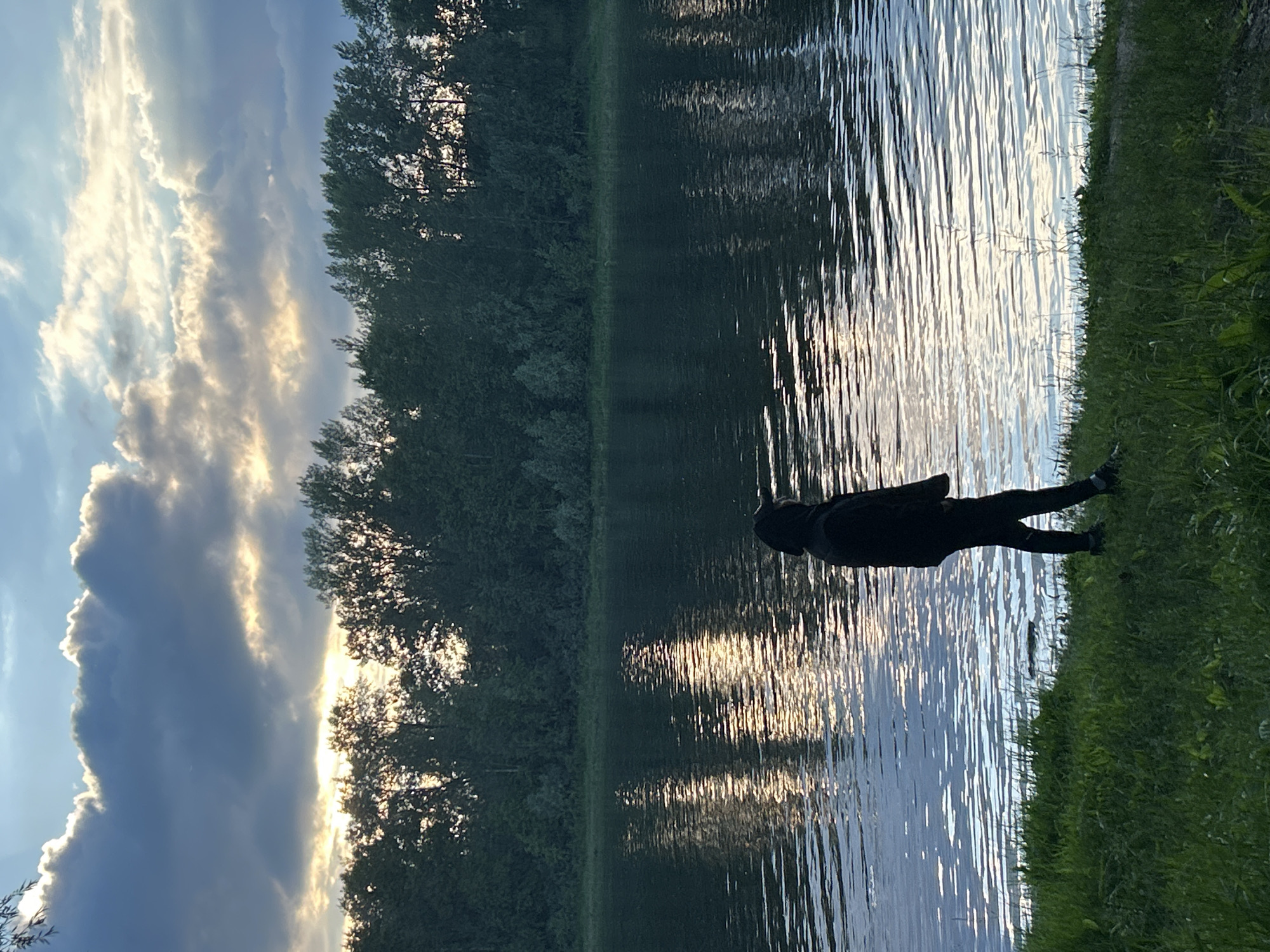 A person in dark clothing lies on their stomach in the grass on the bank of a body of water. Their legs are extended and their head is turned towards the water. The water is calm with ripples on its surface, reflecting the sky and trees. The sky is partly cloudy with streaks of sunlight breaking through the clouds. The sun appears to be low in the sky, casting a warm glow on the scene, suggesting either sunrise or sunset. The trees on the opposite bank are dense and green, forming a dark line against the bright sky. The grass on the bank where the person is lying is vibrant green. The overall mood is peaceful and serene.