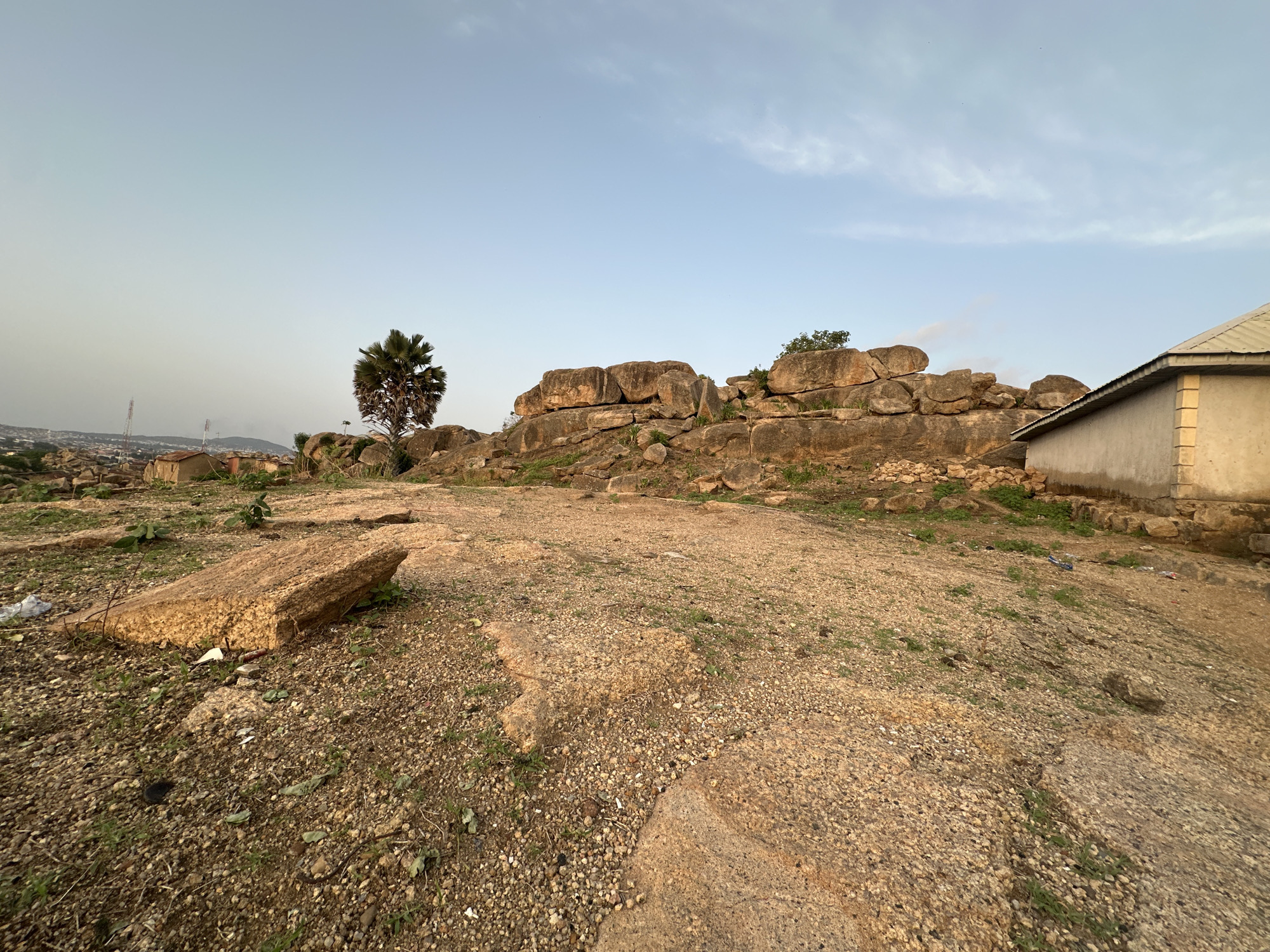The image shows a rocky landscape in Jos, Nigeria, with a clear sky overhead. In the foreground, there's a rugged, uneven terrain covered in small rocks and sparse vegetation. A large, rectangular rock sits prominently on the ground. Further back, a formation of large boulders rises, interspersed with trees and greenery. Adjacent to the rocks, a simple, single-story building with a visible roof and plain, light-colored walls is situated. In the distance, a cityscape is visible, indicating an urban setting. There are no people visible in the image.