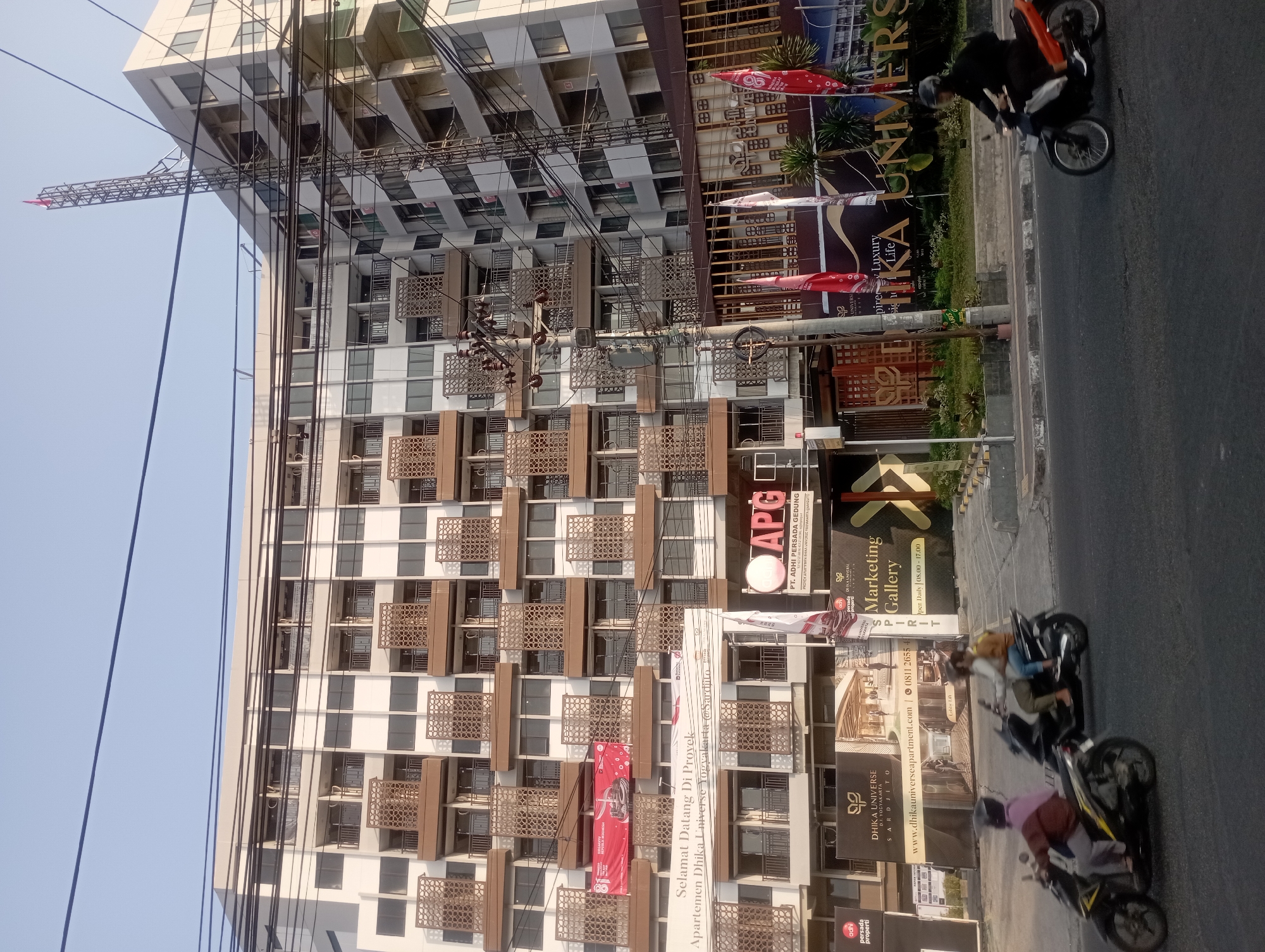 This is a street-level, eye-level, medium shot of the exterior of the Dhika Universe Apartment complex in Yogyakarta, Indonesia. The building is a large, multi-story structure with a white and brown facade, featuring numerous balconies with decorative screens. A large banner that reads "SELAMAT DATANG DI PROYEK Apartemen Dhika Universe Yogyakarta @Sardjito" is prominently displayed on the building. Several red flags are also visible. In the foreground, there are three motorcycles in motion on the street. The leftmost motorcycle carries a single rider wearing a pink headscarf and a brown jacket. The middle motorcycle has two riders, one in a yellow top and the other in a dark top and olive pants. The rightmost motorcycle is ridden by a person in black attire. The environment appears to be an urban street with power lines crisscrossing overhead, a utility pole with transformers in the middle ground, and various advertisements and signage related to the apartment complex. The sky is clear and bright, indicating daytime.