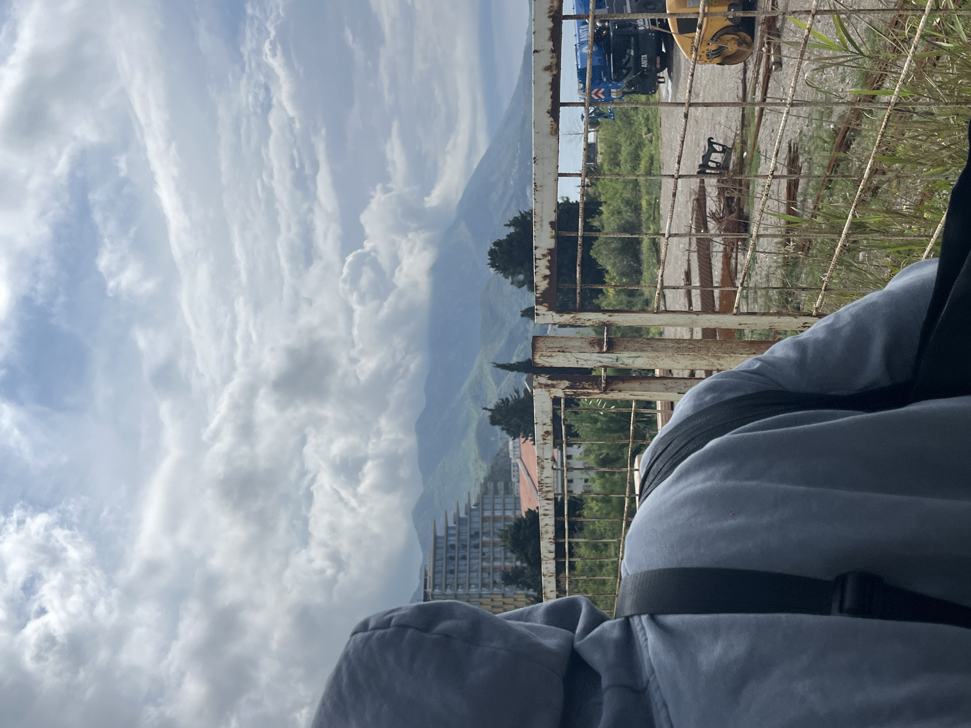 The image captures a scenic view from a slightly elevated position, featuring a person in the foreground observing a landscape through a rusty metal fence. The person, seen from the back, wears a light grey or blue hooded top with a dark strap, possibly from a backpack, visible over their right shoulder.
The scene unfolds with a vast expanse of mountains in the background, partially veiled by large, white, streaky clouds under a bright, partially cloudy sky. Below the mountain range, several buildings are visible, including a modern-looking structure with multiple levels and glass windows.
In the middle ground, behind the fence and to the right, there are signs of industrial activity or construction. A yellow excavator arm is visible, along with part of a blue crane truck. The text "ANITA" is faintly visible on the side of the crane truck. The ground behind the fence appears to be an unpaved area with some sparse grass and weeds.
The weather appears to be daytime, with significant cloud cover but also visible patches of blue sky, suggesting a partially cloudy day. The rust on the fence indicates age or exposure to elements. The overall setting combines natural mountainous beauty with elements of human development and industry, consistent with a location like Boreti, Montenegro.