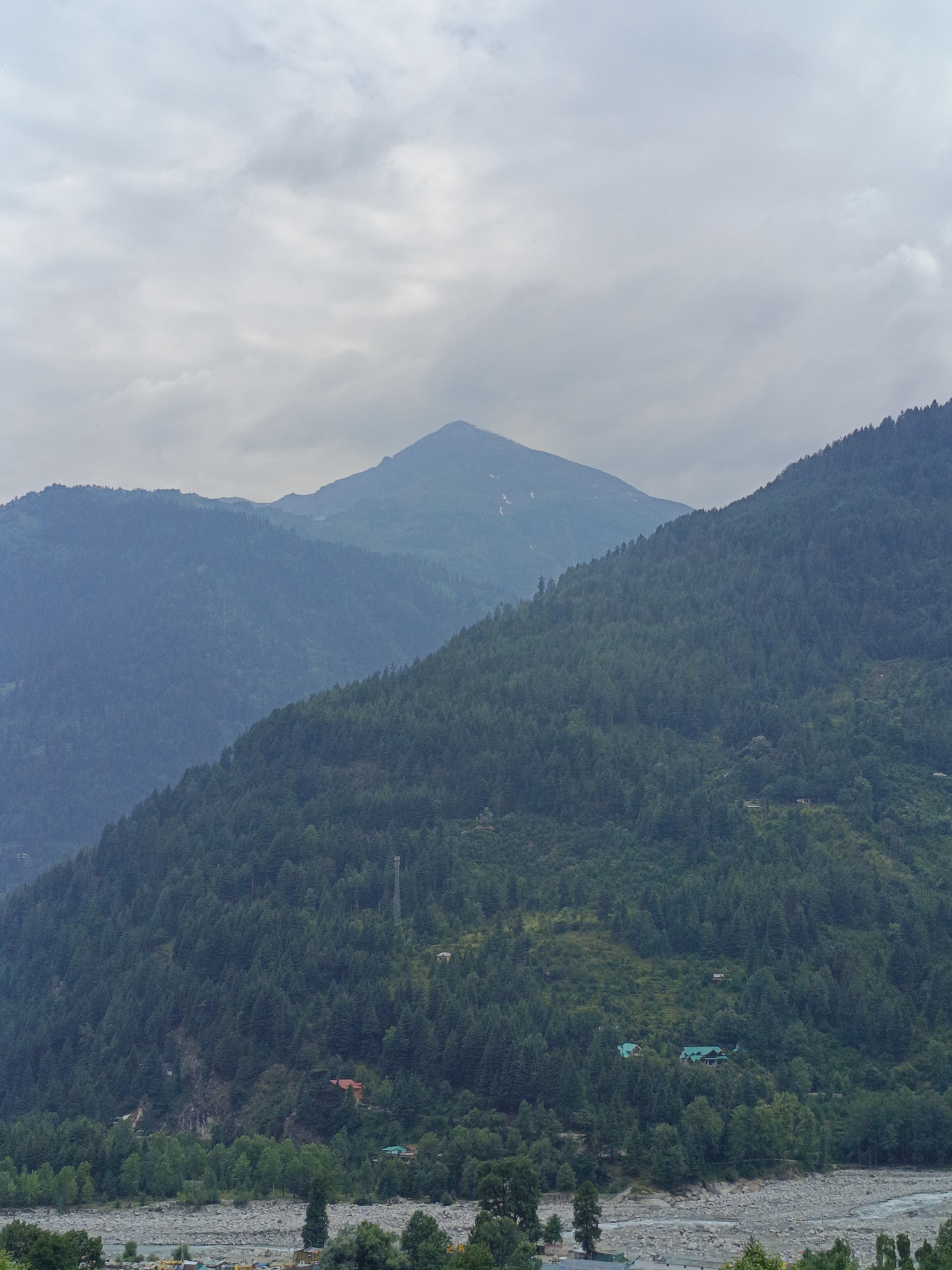 The image depicts a mountainous landscape in Vashisht, India, under a cloudy sky. The foreground features a rocky riverbed with scattered trees and some small structures, possibly houses or guesthouses, nestled amongst them. Beyond the riverbed, a densely forested mountain slope rises steeply, dotted with more scattered structures and a tall communication tower. Further in the background, successive layers of mountains recede into the distance, with the tallest peak in the center appearing lighter due to atmospheric haze. The overall atmosphere is serene and natural, characteristic of a Himalayan region.