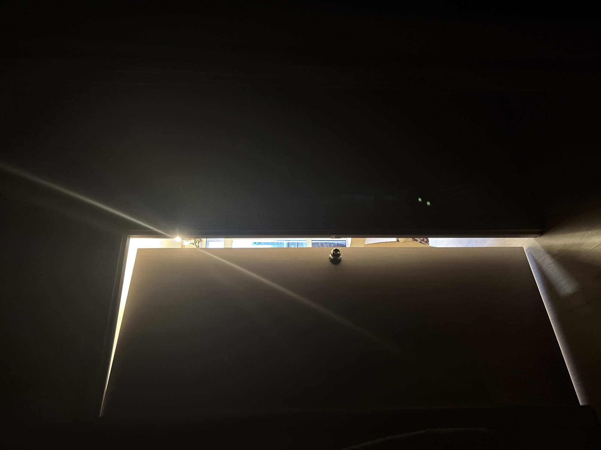 The image shows a partially open door in a dimly lit room. Sunlight streams through the gap, illuminating the door's surface and the floor of the adjacent room. The other room appears to have a window with a view of a city, potentially Perm, Russia, though it is difficult to confirm this location. The door is a solid color with a round knob visible. The scene is a high-contrast interior shot. No people are visible.