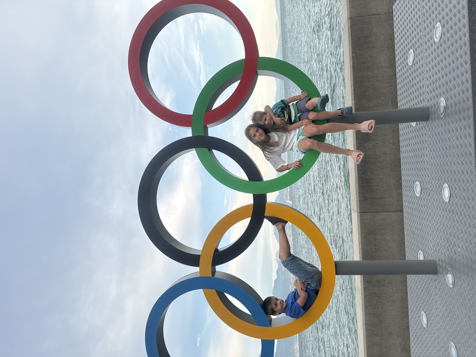 In Lausanne, Switzerland, on the shore of Lake Geneva, five Olympic rings are displayed. On the left, a young boy in a blue t-shirt and denim shorts sits inside the yellow ring, his legs extended. To his right, on the green ring, a teenage girl in a white t-shirt and turquoise shorts sits with her arm around a younger boy wearing a striped blue and green t-shirt and blue shorts. They are all positioned on the lower part of the rings, which are supported by grey poles. The Olympic rings are set against a backdrop of the lake, with mountains visible in the distance under a cloudy sky. The ground in the foreground is a diamond-patterned metal surface with several embedded circular lights.