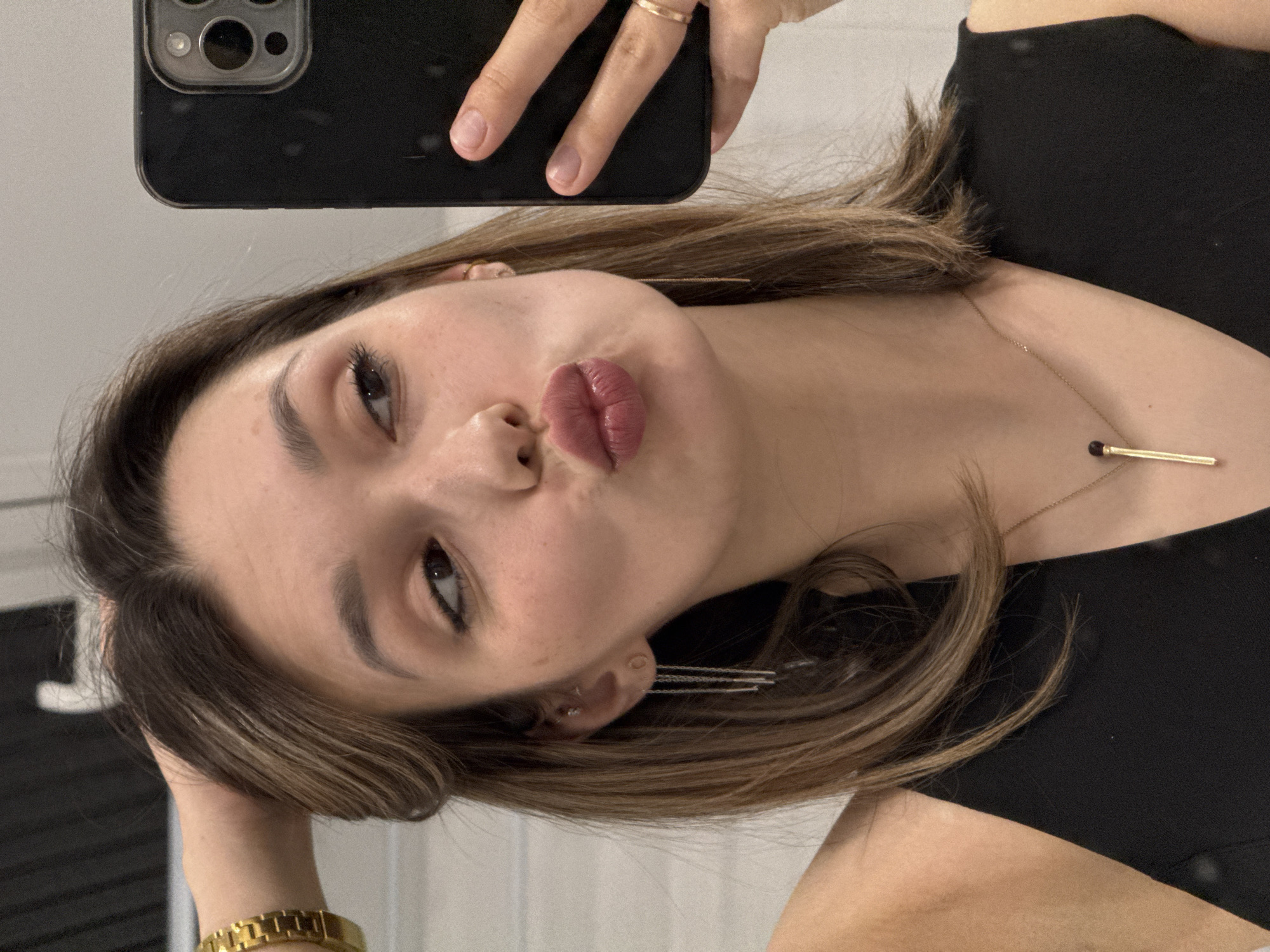 A woman is taking a selfie with a smartphone. She has brown hair and is wearing a black top and gold jewelry. The background is indistinct.