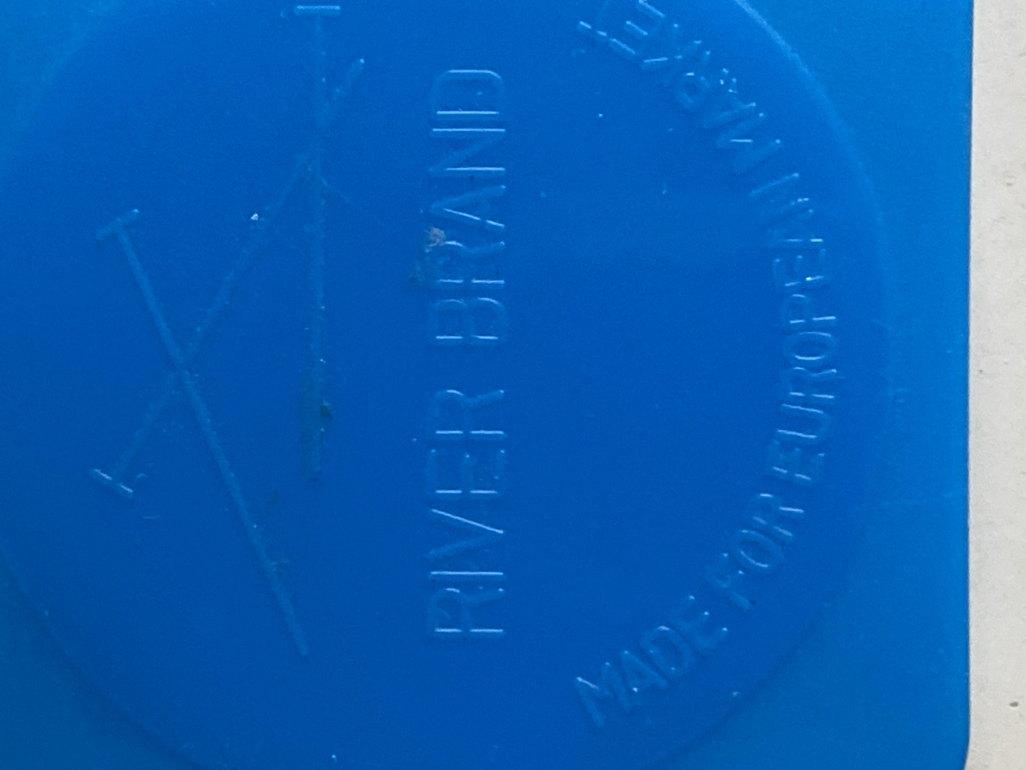 The image is a close-up of embossed text on a blue surface. The text reads "RIVER BRAND" in the center, and in a circular pattern around the bottom, it reads "MADE FOR EUROPEAN MARKE". There is also a faint outline of two crossed lines, possibly representing oars or a similar symbol, above the "RIVER BRAND" text. The blue surface appears to be a plastic material. The image does not contain any information or visual cues that would allow for its location to be determined in the city of Jauro Yinu, Nigeria, or any other specific location.
