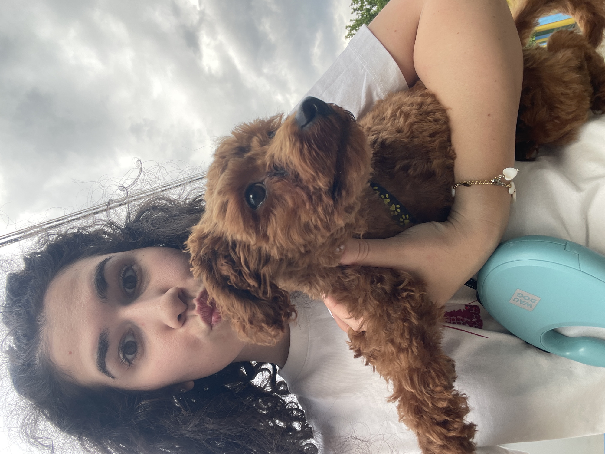 A person with dark curly hair and dark eyes is holding a brown dog with curly fur. The person is wearing a white shirt and has a gold bracelet with white charms on their wrist. They are making a kissing face. The dog has a black collar with yellow paw prints on it. Next to the person's arm is a blue dog leash handle. The background is a cloudy sky. The location of the photo is not possible to determine in the city of Sofia, Bulgaria.