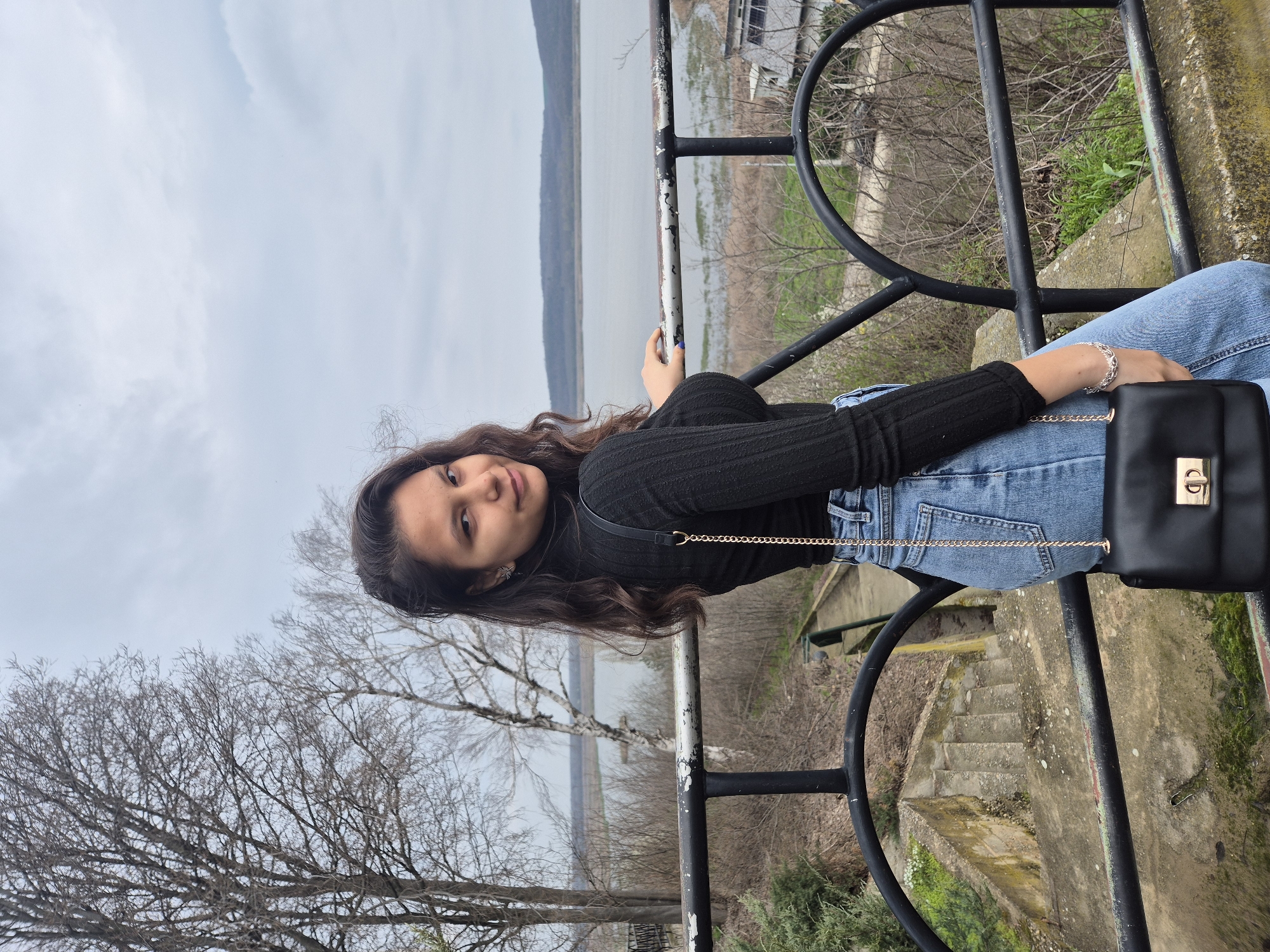A young woman is posing for a photo in what appears to be Srebarna, Bulgaria. She is leaning against a black metal railing, with a body of water and a landscape visible in the background. She is wearing a black turtleneck sweater, blue jeans, and a black purse with a gold chain and clasp. She has long brown hair and is wearing a bracelet on her left wrist. The railing shows signs of wear and age. Bare trees and vegetation are visible in the surrounding environment. The sky is overcast. Steps lead down from behind the railing. Buildings can be seen on the opposite shore in the background.