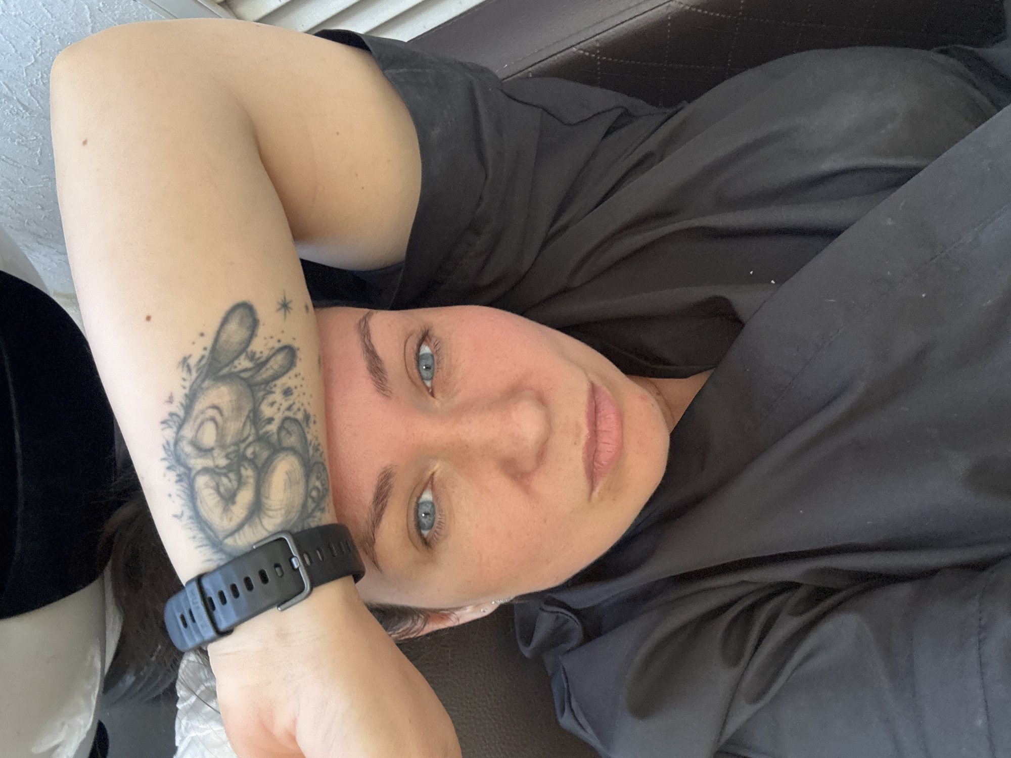 A light-skinned individual with blue eyes is lying down and facing the camera. The person has a tattoo of a rabbit on their left forearm and is wearing a black wrist watch. They are wearing a black shirt and lying on what appears to be a dark brown cushioned surface. No information indicates that the location is the city of Chaikovsky, Russia.