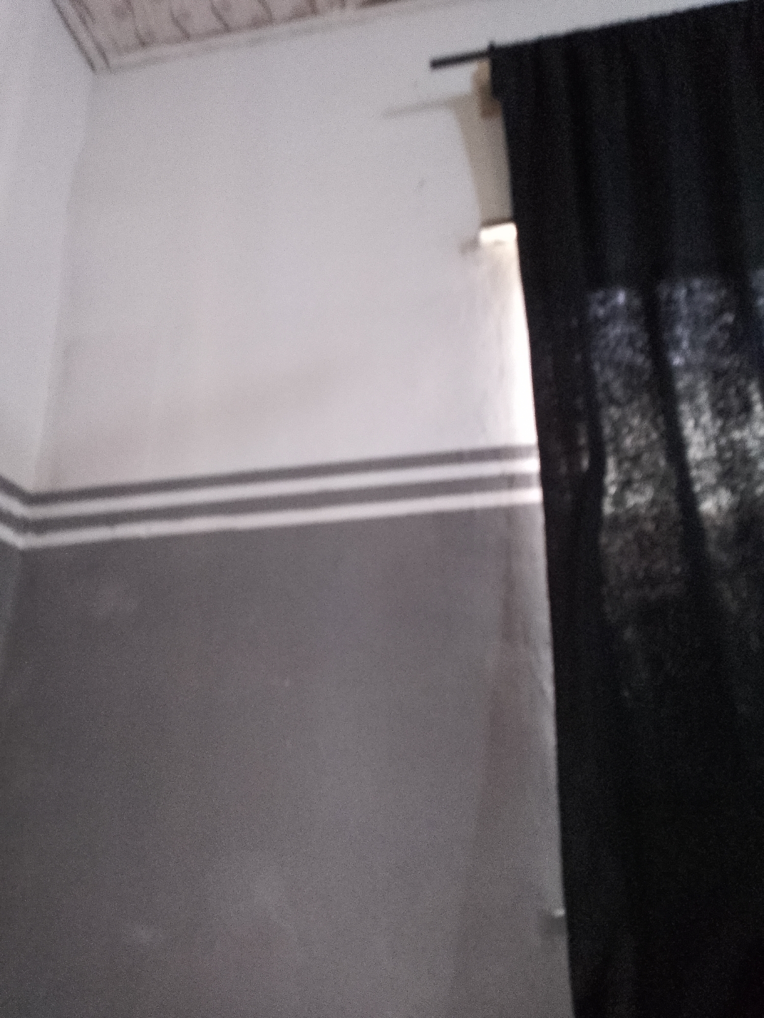 The image shows an interior scene, likely a room, with a distinct wall design. The wall is painted in two horizontal sections: the upper portion is white, and the lower portion is gray. A thin band of two parallel, lighter gray lines separates the white and gray sections. A black curtain is partially visible on the right side of the image, drawn across a window. The ceiling, visible in the top left corner, appears to be covered with some form of paneling. The lighting suggests an indoor environment, possibly with light coming from the window behind the curtain. The scene does not contain any individuals or identifiable objects that would place it specifically within Jalingo, Nigeria.