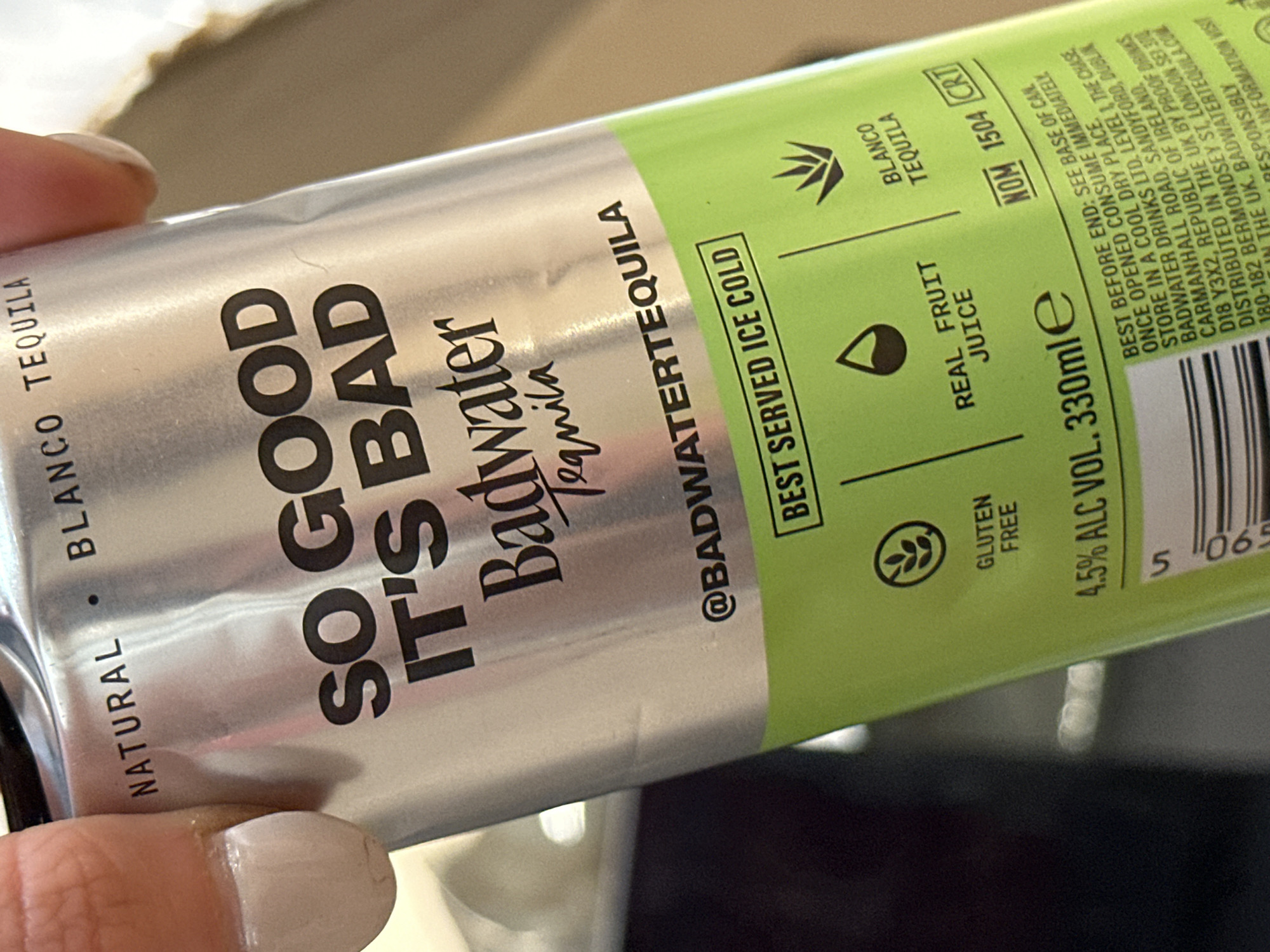 A close-up, high-angle shot depicts a person's hand, exhibiting manicured fingernails with light neutral polish, gripping a silver aluminum beverage can. The can's upper half is silver, transitioning to a lime green band on the lower section. Prominently displayed in black lettering are the words "SO GOOD IT'S BAD" above "Badwater Tequila." Further text includes "NATURAL • BLANCO TEQUILA," "@BADWATERTEQUILA," and "BEST SERVED ICE COLD."
The green band features icons and text indicating "GLUTEN FREE," "REAL FRUIT JUICE," and "BLANCO TEQUILA." Below this, "4.5% ALC VOL. 330mle" is printed, along with "NOM 1504 CRT." The label details storage instructions and distribution information, identifying "PROOF DRINKS, 180-182 BERMONDSEY ST, LONDON SE1 3TQ" as the UK distributor. The can also states "MADE IN THE UK." The background is out of focus, showing indistinct light and dark areas consistent with an indoor environment. The scene is directly linked to Bermondsey Street, London SE1 3TQ, United Kingdom, by the distributor's address on the product label.
