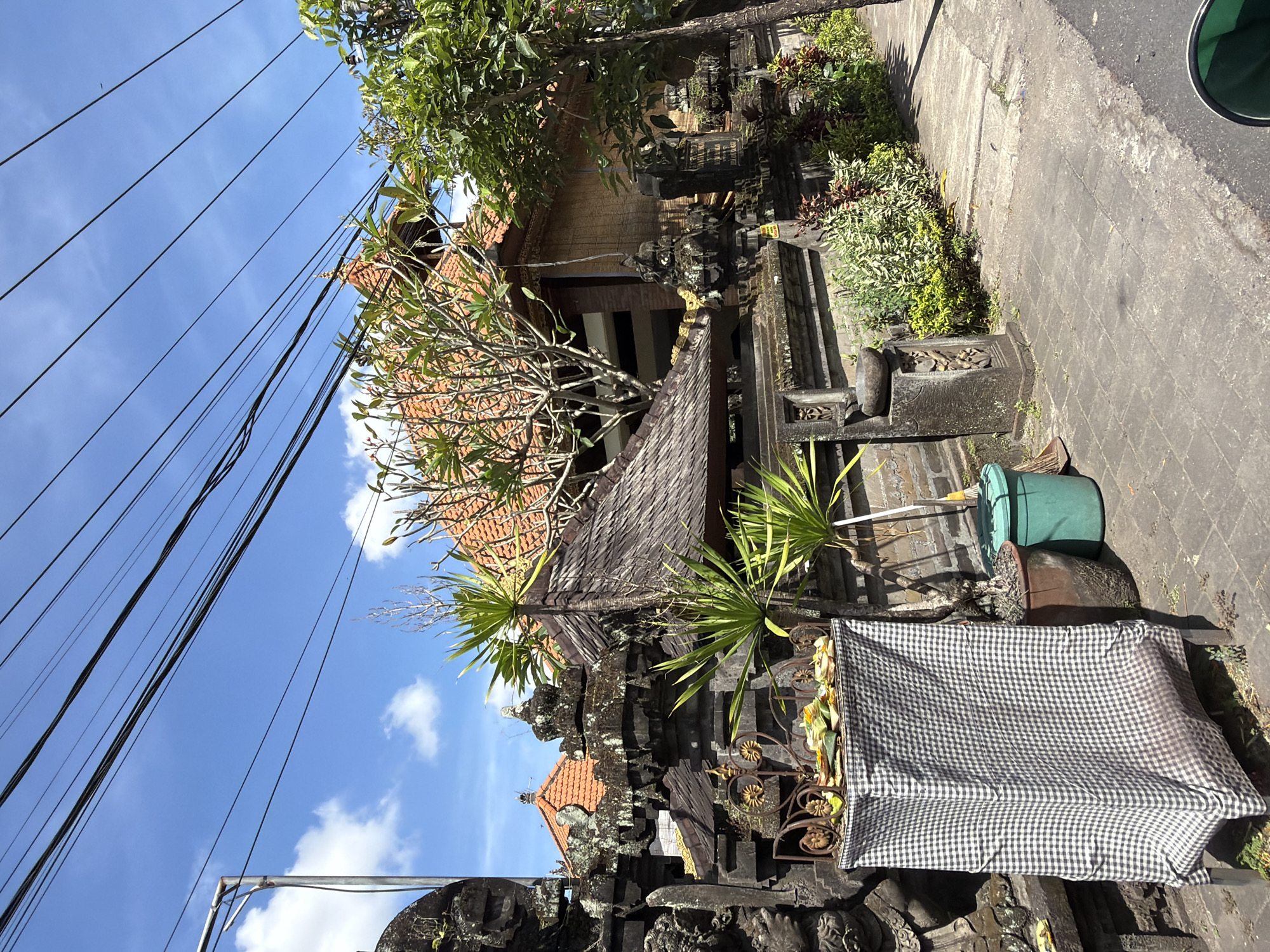 The image captures a scene in North Kuta, Indonesia, showcasing a traditional Balinese architectural style. The foreground features a stone wall adorned with intricate carvings, and a black and white checkered cloth draped over a structure. Behind this, two potted plants stand near a dark green bin. The architecture displays roofs with orange tiles and dark, weathered wood. Overhead, power lines stretch across a blue sky with scattered clouds. A sidewalk is visible on the right side of the frame. The composition is in a slightly low angle.