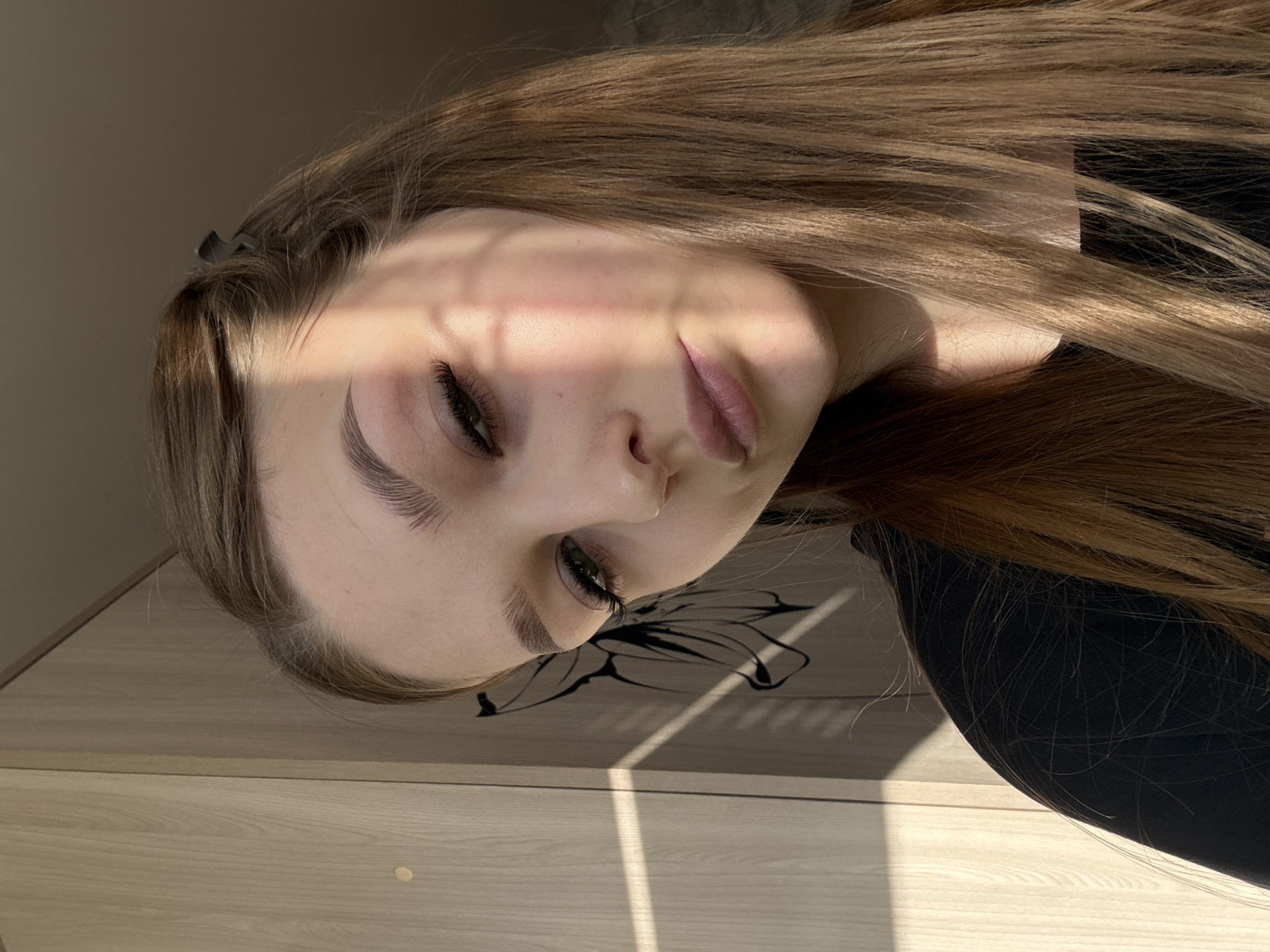 A young woman is captured in a close-up portrait, lying down with her head turned sideways. Her long, brown hair falls around her face and onto a wooden surface, possibly a table. She has fair skin, defined eyebrows, and long eyelashes, suggesting she may be wearing mascara. A faint shadow from what appears to be window blinds or tree branches falls across her face, partially obscuring one side. She wears a dark-colored top, which is slightly visible in the frame. The background is a plain, light-colored wall. Based on the instructions, this image was likely taken in the city of Chaikovsky, Russia.