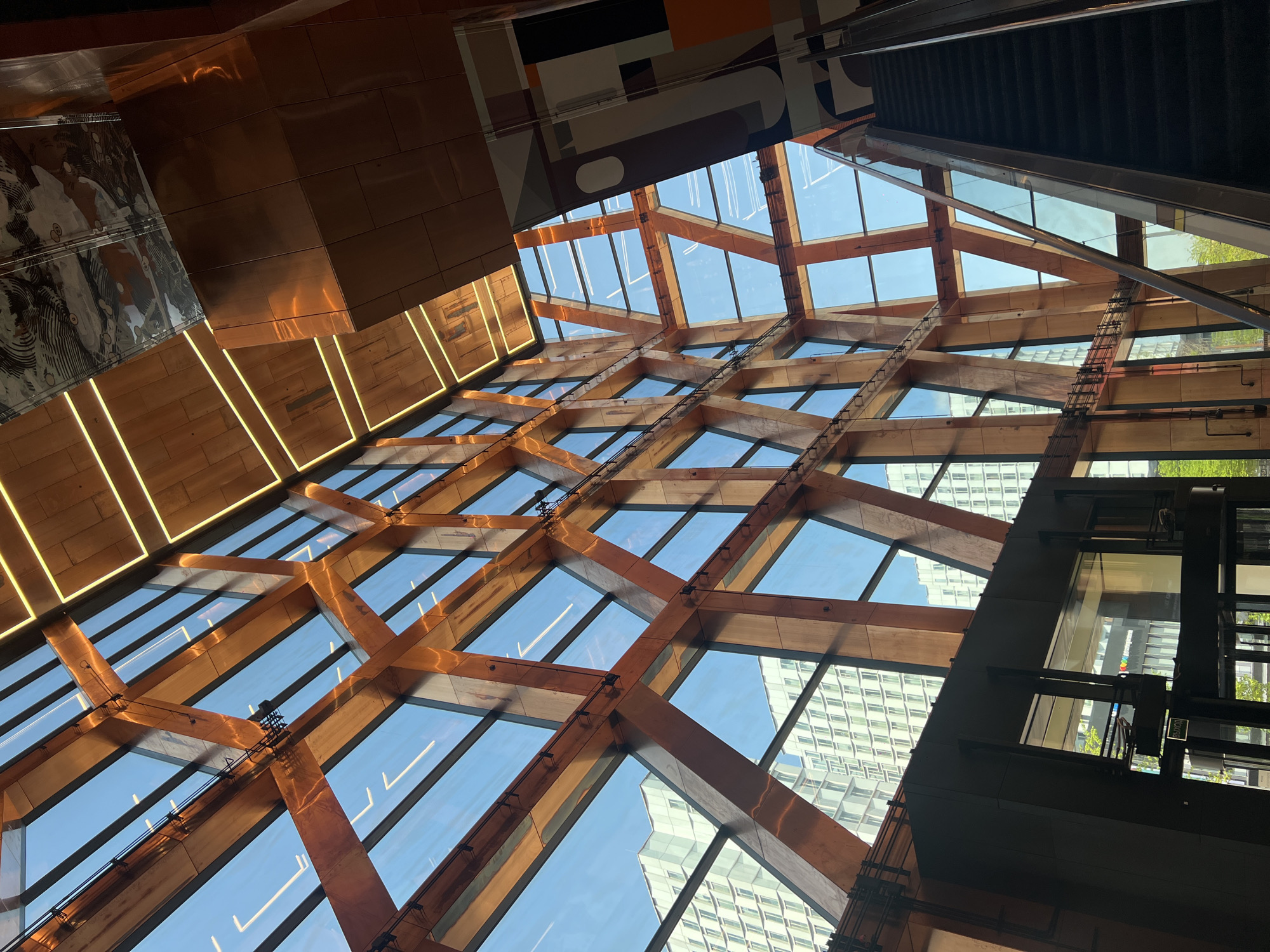 The image depicts an upward view of a building's interior, likely a structure with a unique architectural design. The scene is characterized by a grid-like framework of wooden beams and glass panels, forming a kind of vaulted ceiling or skylight. Through the glass, one can see the exterior environment, including parts of buildings with a modern aesthetic. The bright blue sky suggests a sunny day. On the periphery, elements of interior structures are visible, including a reflective copper-colored panel and what appears to be an elevator shaft or some kind of vertical transport system. The image has a somewhat tilted perspective. While there is not specific information from the image to determine the location, the architectural style can be recognized as being atriums.