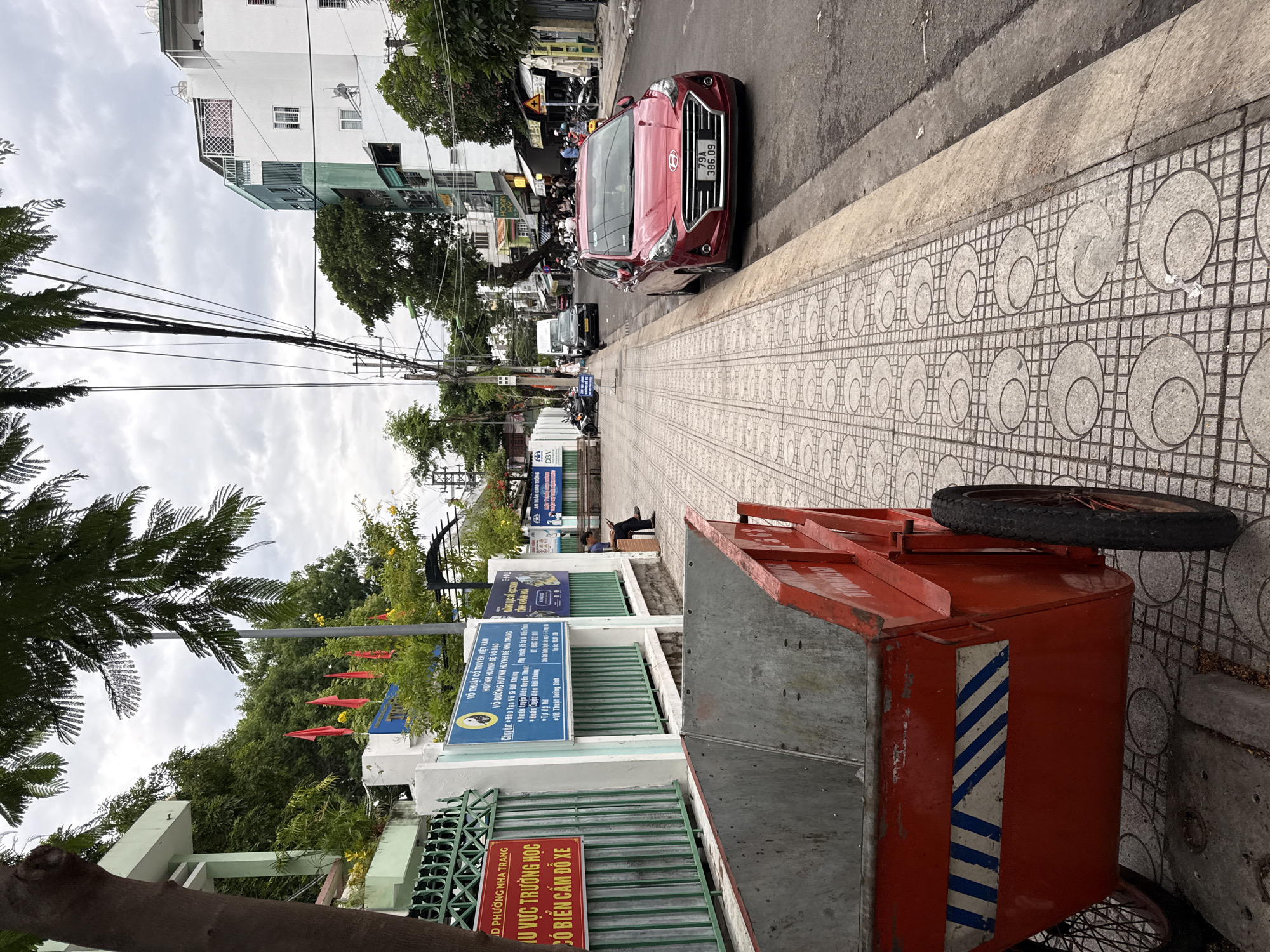 A red cart with two wheels sits on a patterned sidewalk. Behind the cart, a building with green trim and fences is visible, displaying several red flags and signs. Further down the street, a red car is parked on the right side of the road. The street is lined with buildings, trees, and utility poles with numerous wires. Several people are seen on the sidewalk and in the street, some riding motorcycles. The sky is overcast. The signs indicate the location is Võ Thuật Truyền Thống Huỳnh Đệ and likely in Nha Trang, Vietnam. One sign on the green fence also mentions "TU VỰC TRƯỜNG HỌC CÓ BIỂN CẤM ĐỖ XE", which translates to "school area, no parking". Another sign further down mentions "TOÀN GIAO THÔNG", indicating traffic safety.