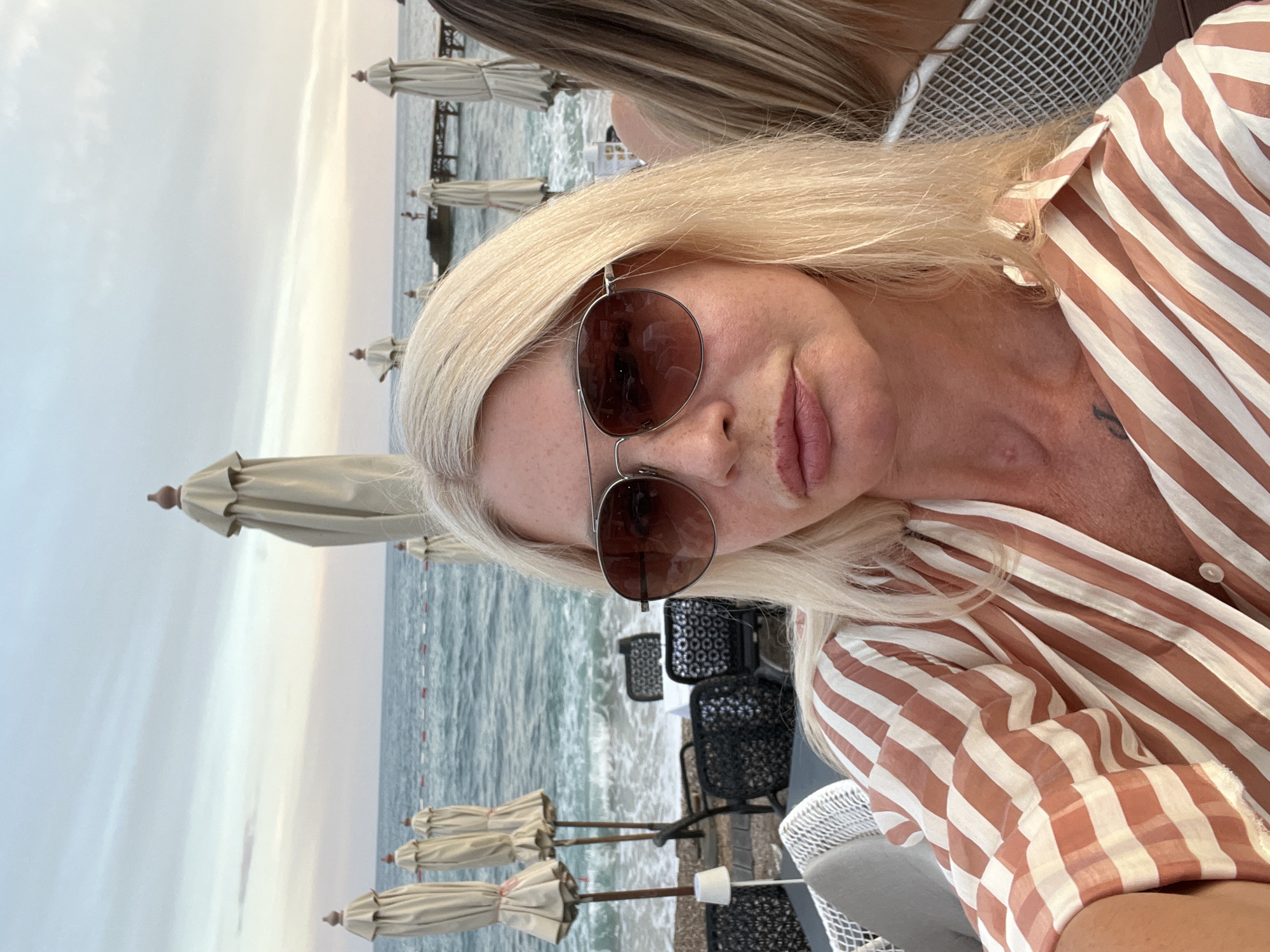 The image shows a close-up of a woman with blonde hair and sunglasses, wearing a striped shirt. She is seated at an outdoor table near a beach. In the background, there are closed beach umbrellas, chairs, tables, and the ocean. The sky is overcast with hints of pink and orange, suggesting it is either sunrise or sunset. There are other people and beach structures visible in the distance.