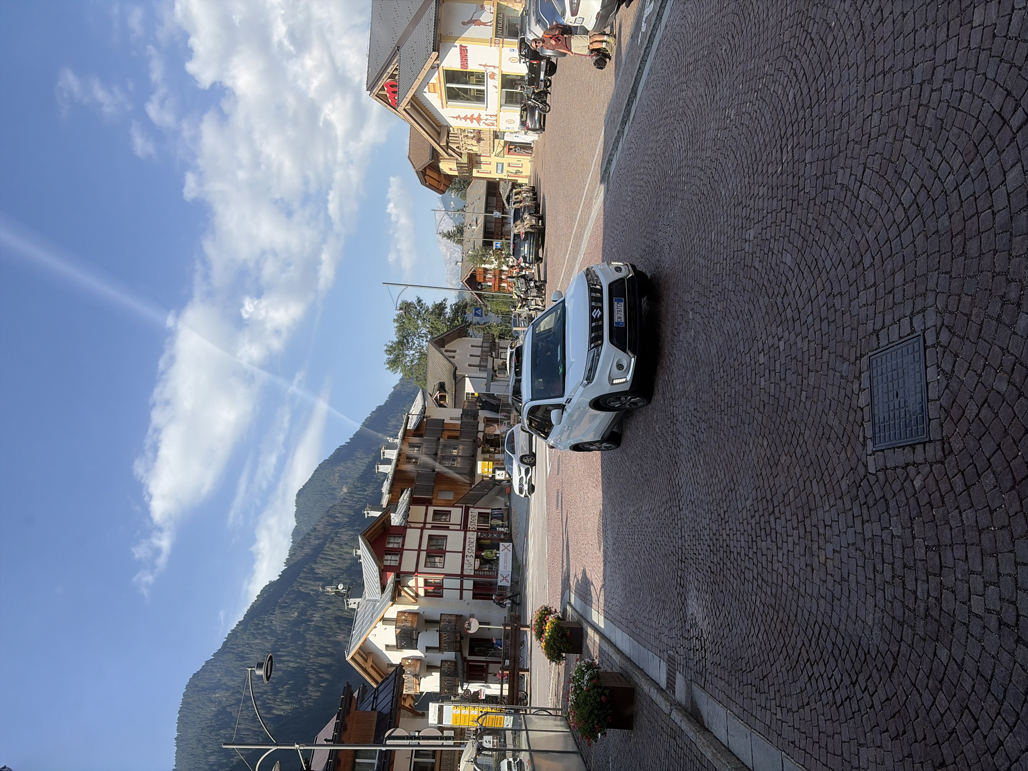 The image captures a street scene in Campitello di Fassa, Italy, under a bright, partly cloudy sky. The foreground is dominated by a cobblestone street with a curved pattern. A white Suzuki Vitara is parked in the middle of the street, facing towards the viewer. To the left of the Vitara, a row of buildings with traditional alpine architecture lines the street. These buildings feature wooden balconies, red and white facades, and grey roofs. Flower boxes with colorful blooms are visible in front of some of the buildings.
Further down the street, more buildings are visible, some with shops on the ground floor. A sign for "SPORT 365" is prominent on one of these buildings. Several other cars and motorcycles are parked along the street or in motion. A person with a dog is standing on the right side of the street near a parked car. In the background, a majestic mountain covered in pine trees rises above the town, with its peak partially obscured by clouds. The overall impression is a bustling, picturesque mountain town on a sunny day.