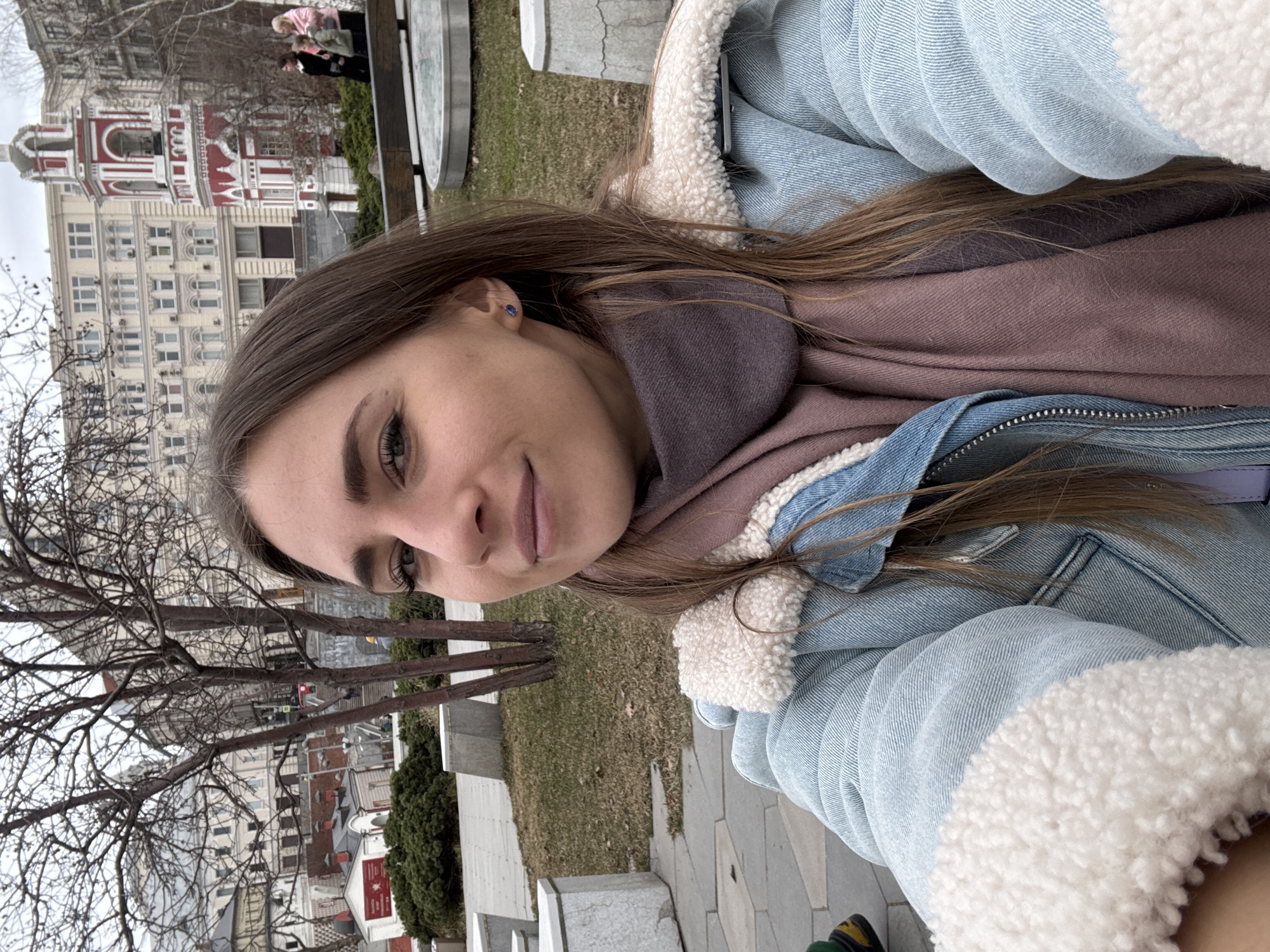 A young woman is featured in a close-up, selfie-style photograph, positioned centrally in the foreground. She has long, straight brown hair, light skin, and is looking towards the viewer with a slight smile. She wears a light blue denim jacket with cream-colored sherpa lining on the collar and cuffs, and a dark mauve scarf wrapped around her neck. A small, blue stud earring is visible in her right ear.
The background depicts an urban park environment in Moscow, Russia. The most prominent identifiable structure is the red and white bell tower of the Church of the Nativity of the Theotokos at Putinki, clearly visible in the middle ground. This church is located in Pushkin Square. Behind and to the left of the bell tower, a multi-story building with a light-colored facade and numerous windows is present. Further to the left, a smaller, reddish building with white trim is partially visible down a slight incline.
The park features bare deciduous trees, areas of sparse, dry grass, paved walking surfaces, and light-colored concrete seating or planters. Low green shrubbery is also evident. In the middle ground, to the right of the bell tower, three figures are observed: a woman in a dark coat, and another woman in a light pink coat with an adjacent child. The sky is overcast, indicating a cool, possibly late autumn or early spring day.