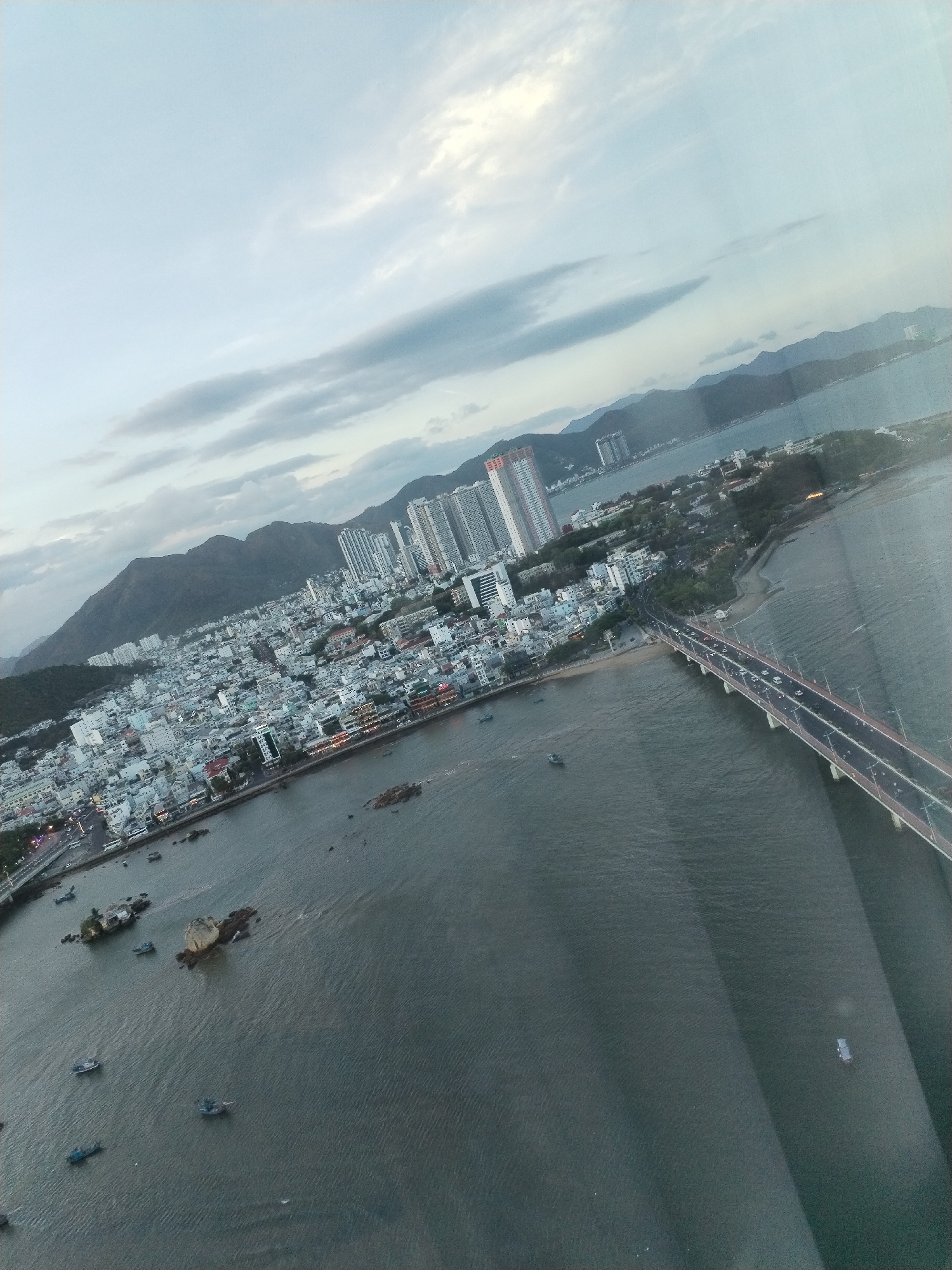Aerial view of the coastal city of Nha Trang, Vietnam. The cityscape is a mix of high-rise buildings and low-rise structures situated between mountains and the coast. A large bridge spans a body of water, carrying vehicular traffic. Numerous small boats are visible on the water. The sky is overcast.