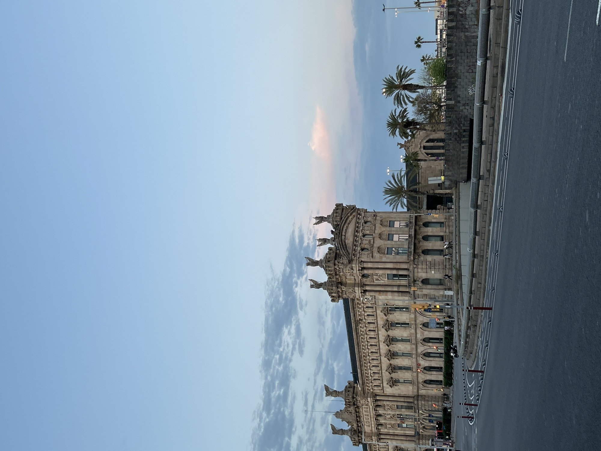The image depicts a serene urban scene, likely captured during twilight (either dawn or dusk), showcasing a grand architectural building alongside a quiet city street.
The primary subject is an ornate, classical-style building that runs along the left side of the frame. Its facade is richly detailed with arched windows, columns, and intricate stonework. Figures, possibly statues, adorn the roofline and upper ledges of the building. Notably, the word "ADUANA" (Custom House) is clearly visible etched into the building's facade, confirming its identity as a historical Custom House. A few indistinct human figures can be seen on the building's balconies or ledges.
To the right of the building, a multi-lane road stretches, appearing relatively empty. It features distinct white lane markings, red and white traffic bollards, and a metal guardrail separating the lanes from a raised section or pavement. Some motorcycles or scooters are parked or moving slowly on the road. In the background, behind a stone wall, several tall palm trees are visible, characteristic of a Mediterranean climate. Streetlights are present, some illuminated, further supporting the twilight setting.
The sky, occupying the upper portion of the image, transitions from a clear, light blue to lighter tones towards the horizon, interspersed with soft, wispy clouds that show faint hints of pale pink and grey, reinforcing the early morning or late evening atmosphere. The overall impression is one of calm and tranquility in a grand urban setting.