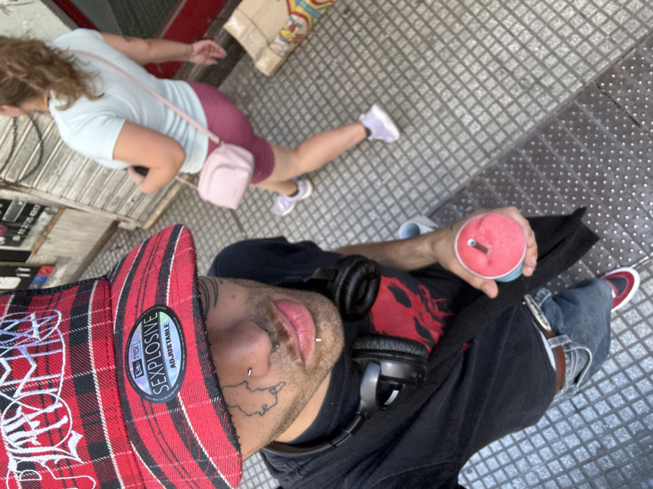 A selfie of a person wearing a red and black plaid hat, black shirt, and headphones around their neck. They have tattoos on their face and are holding a red smoothie with a straw. Another person is walking in the background wearing a light blue shirt, purple shorts, and sneakers. The picture is taken from a high angle on a sidewalk.