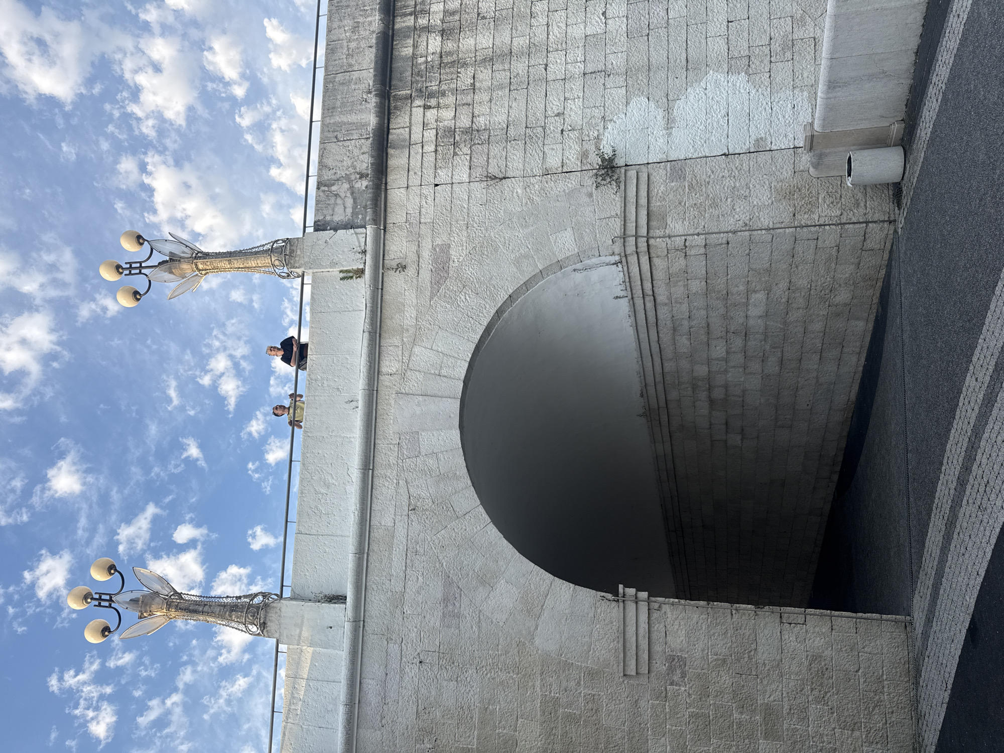 The image captures a view of an archway under a bridge in Sochi, Russia, on a partly cloudy day. The bridge structure is made of white stone blocks. Two street lamps with three globes each are visible on the bridge's parapet. On the bridge, two individuals are standing near the railing. One person, wearing a yellow shirt, is looking up and to their left. The other person, in a black shirt, is leaning on the railing and looking towards their left. The archway leads into a dimly lit tunnel. The foreground shows a paved surface with a pattern of dark asphalt and grey cobblestone. A light grey trash bin is situated to the right of the archway. The sky is blue with scattered white clouds.