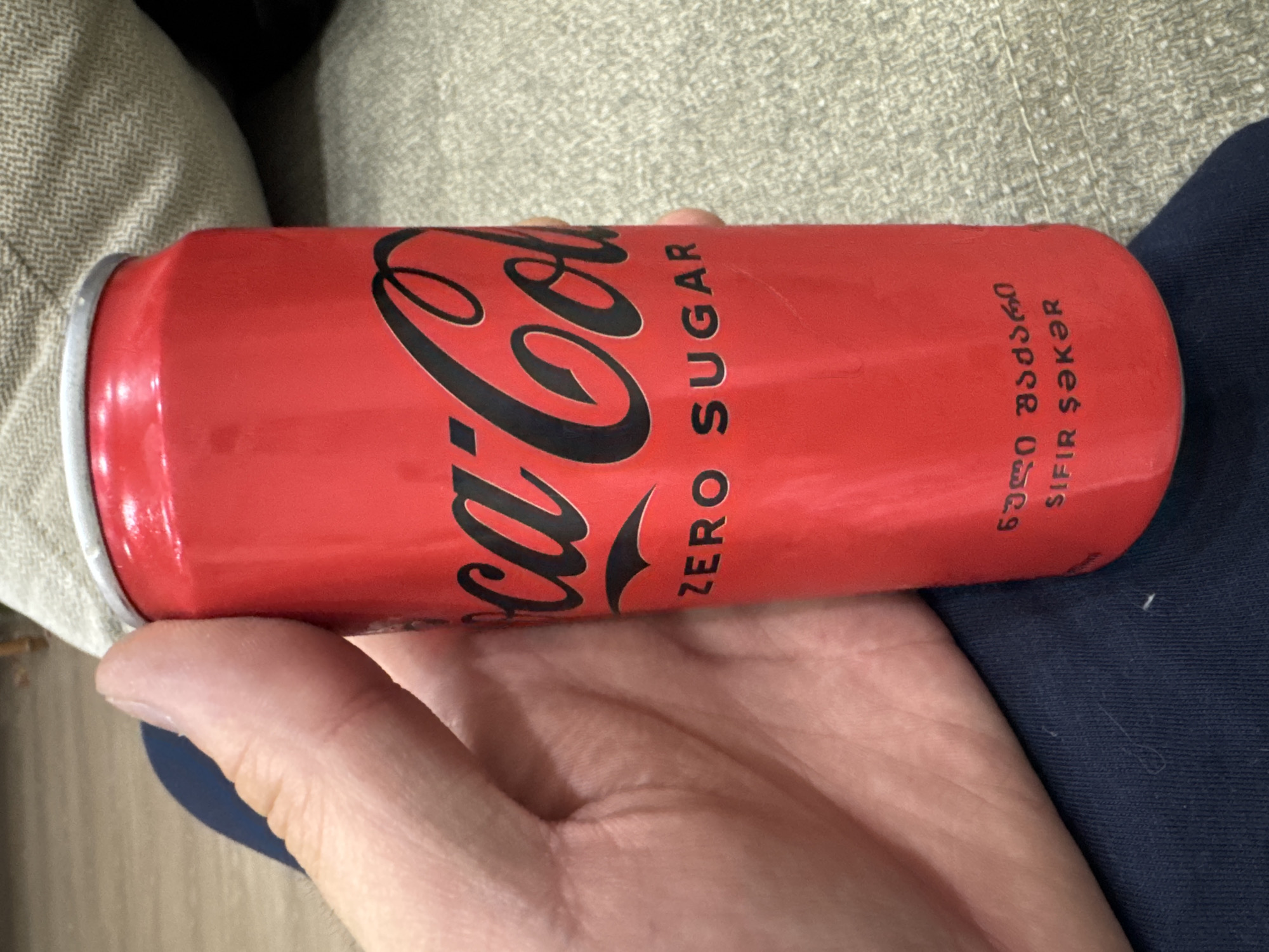 A hand is holding a Coca-Cola Zero Sugar can. The can is red with the Coca-Cola logo in black script and "ZERO SUGAR" in black text. Below that, there is text in Georgian script. The background shows a textured surface, likely a couch or chair, and a blue fabric. The image does not provide any information about the location in the city of Tbilisi, Georgia.