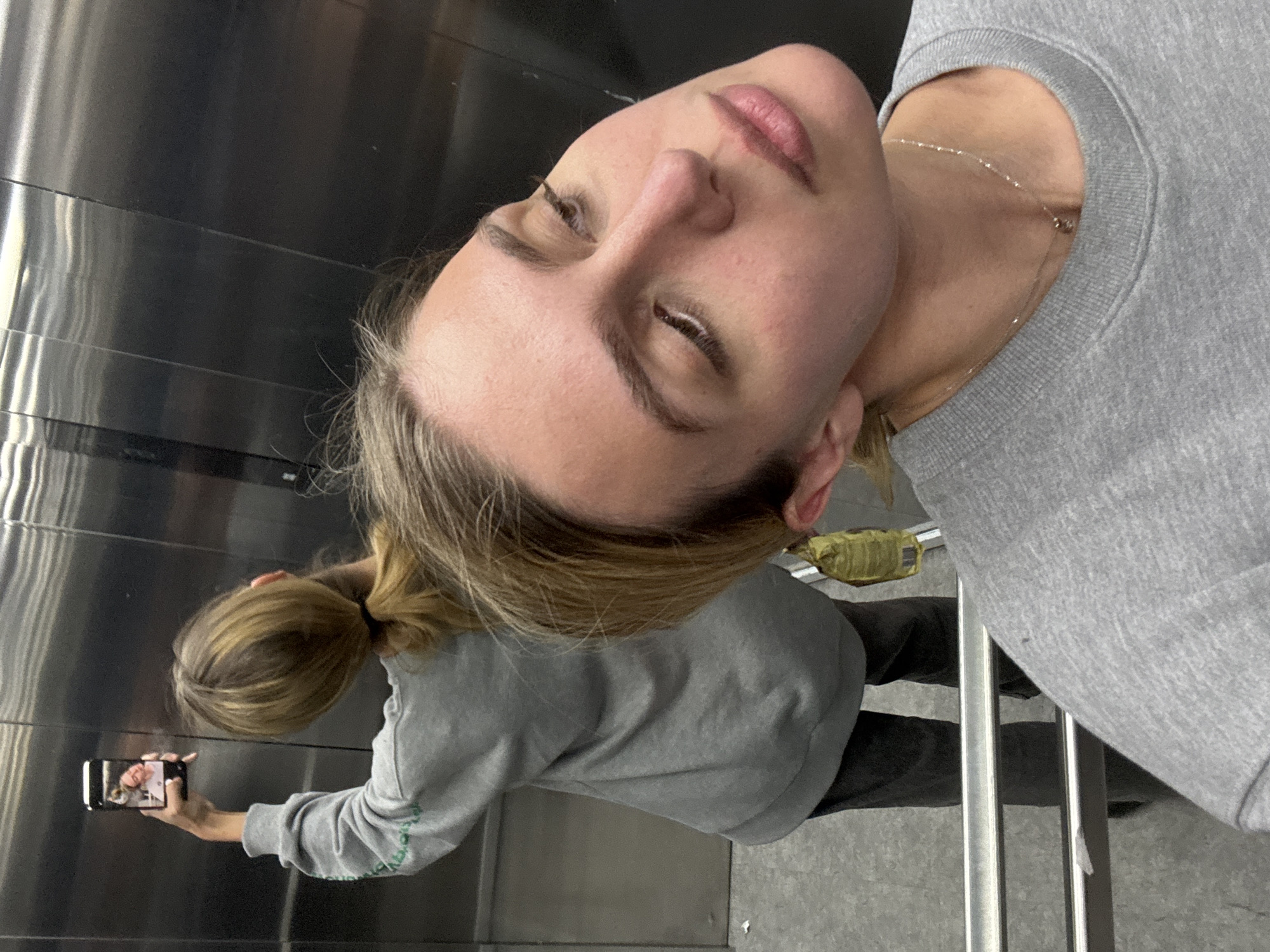 An upward-tilted, close-up selfie captures a woman reflected in a mirror or polished metallic surface, possibly within an elevator or hallway. The woman, with fair skin and light brown hair pulled back into a ponytail, is seen from the chest up. She wears a light gray long-sleeved sweatshirt and a delicate silver chain necklace. Her eyes are partially closed, and her lips are slightly parted.
In the background, her reflection is visible from the waist up. She is holding a black smartphone in her right hand, positioned to capture the selfie. The screen of the phone clearly displays a smiling face, indicating she is in the process of taking the picture. The reflective surface she is facing features horizontal panel lines. To the lower right of the reflection, some vertical metallic bars or railings are visible, and the floor appears to be a gray, textured surface. A small, crumpled yellow object is faintly discernible behind her in the reflection. The scene is illuminated by indoor lighting. No discernible text is visible in the image.