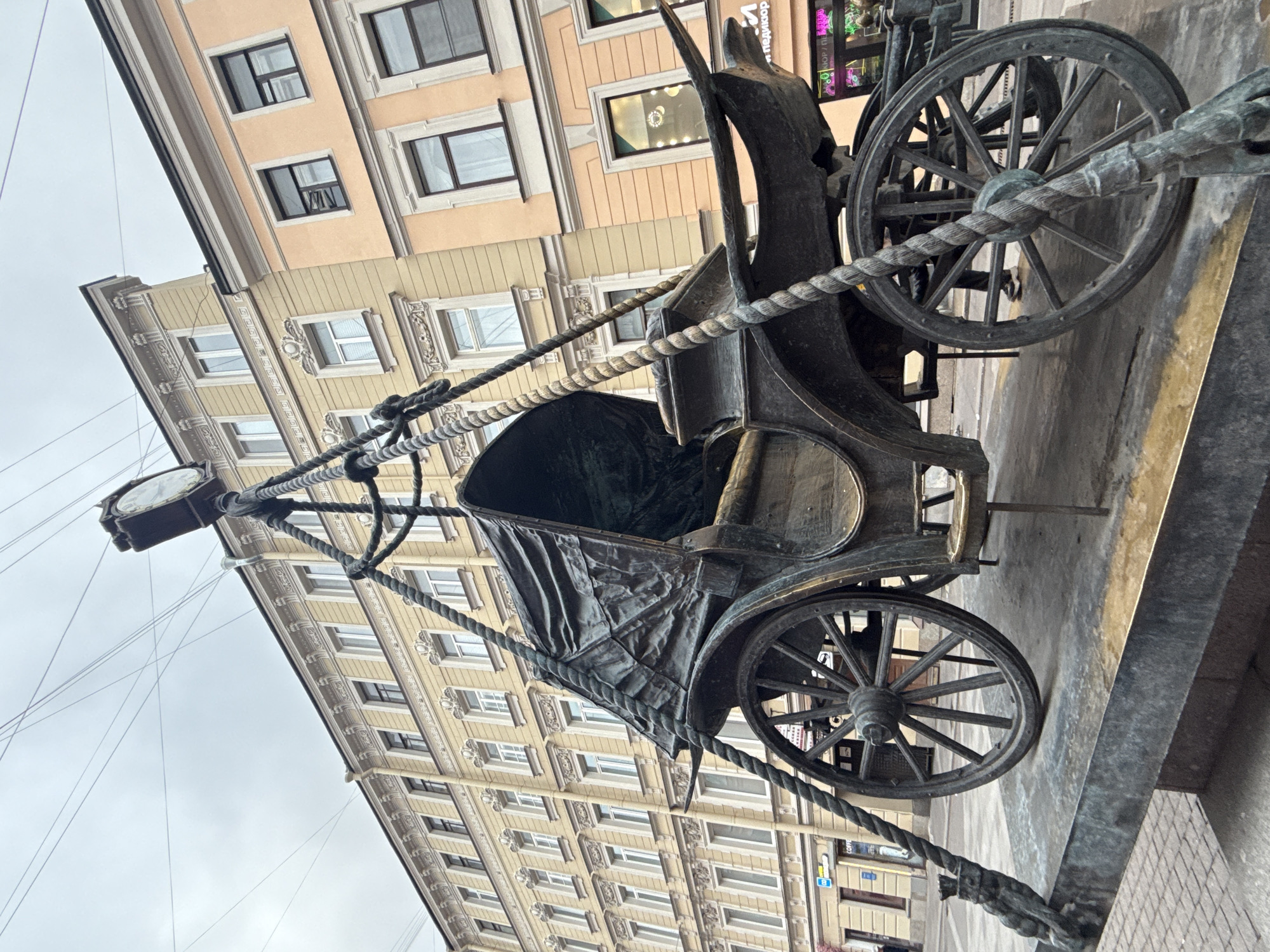The image captures a bronze statue of a historical carriage pulled by horses, located in Saint Petersburg, Russia. The carriage is elaborately detailed with large wheels and a draped covering. It is connected to a lamppost clock via a thick, rope-like bronze structure. The buildings in the background are multi-story, with beige-colored facades and numerous windows, contributing to the city's architectural style. The sky is overcast, suggesting a cloudy day. The statue is positioned on a raised platform with a metal edge. Electrical wires are visible in the sky.