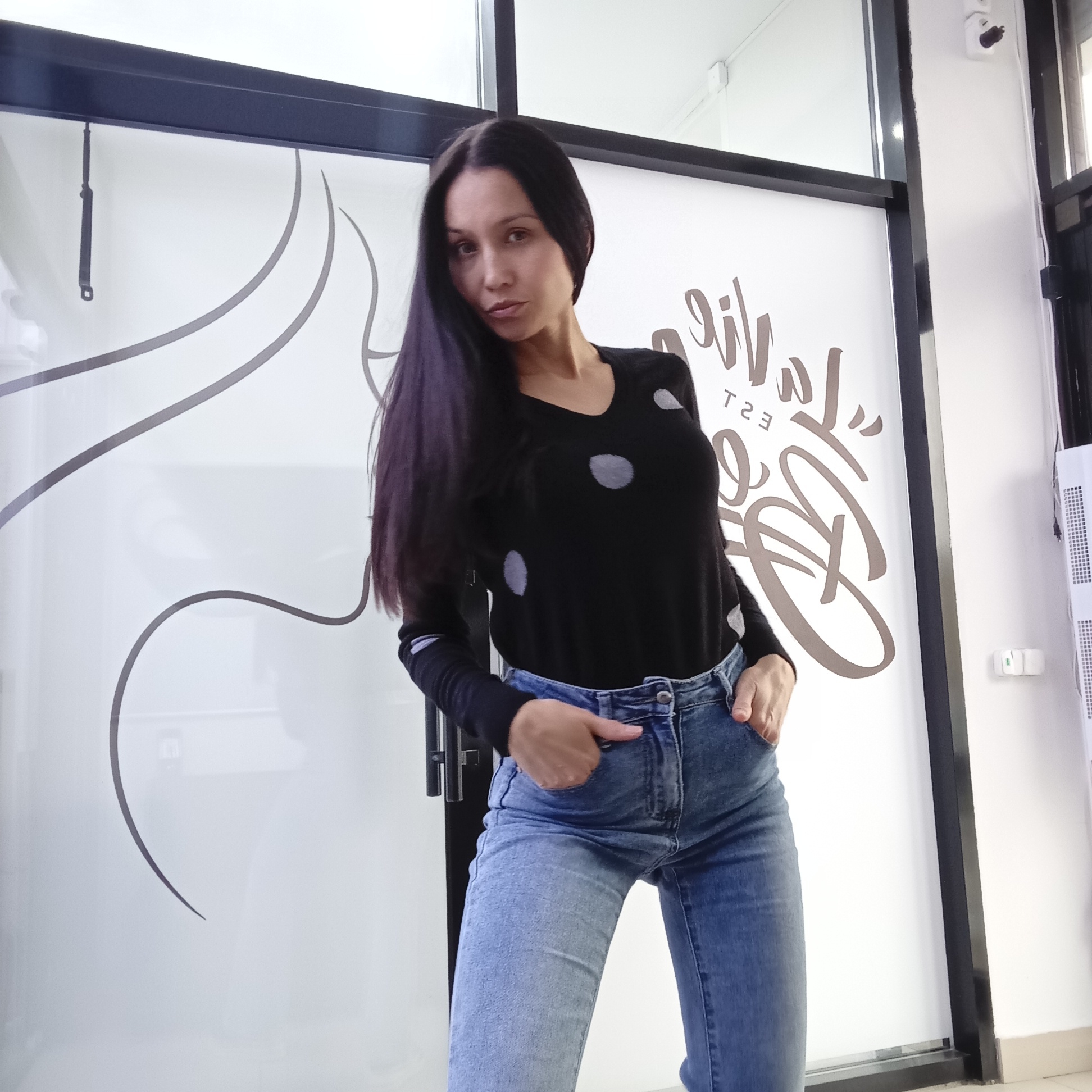 A woman is standing indoors, posing in front of a frosted glass door. She has long, dark hair and is wearing a black long-sleeved sweater with light-colored polka dots and blue jeans. She has her hands in her pockets. The frosted glass door has decorative swirls and a logo/text design. The background consists of white walls. The environment appears to be a commercial space. The location is Izhevsk, Russia.