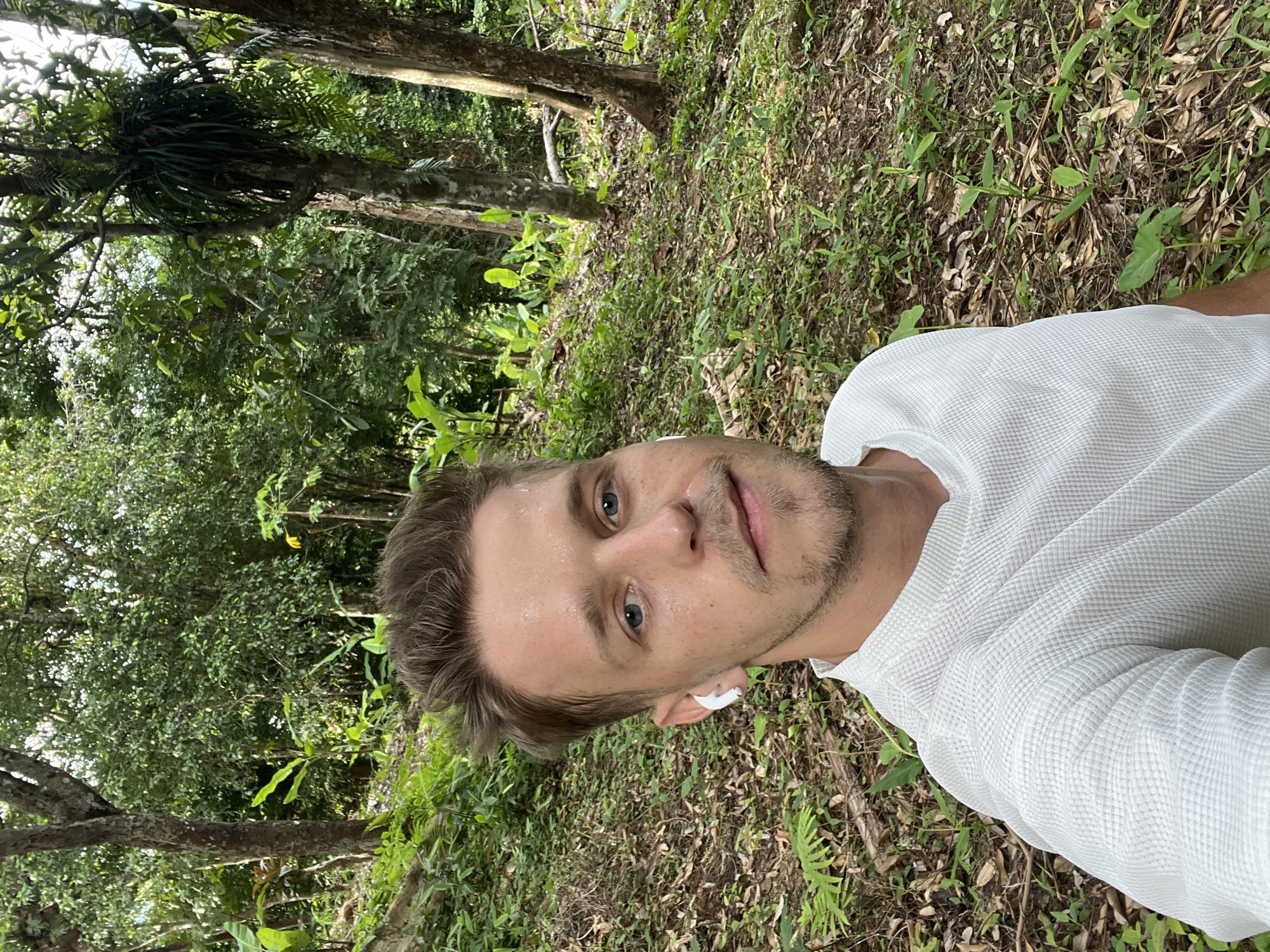 A fair-skinned man with short brown hair and blue eyes is taking a selfie in a lush forest. He is wearing a white waffle-knit shirt and white wireless earbuds. Sweat is visible on his forehead and cheeks. The background is filled with dense green foliage, including trees, large leaves, and brown leaf litter on the ground. The lighting suggests it is daytime, with sunlight filtering through the trees. The location is identified as Choeng Thale, Thailand.